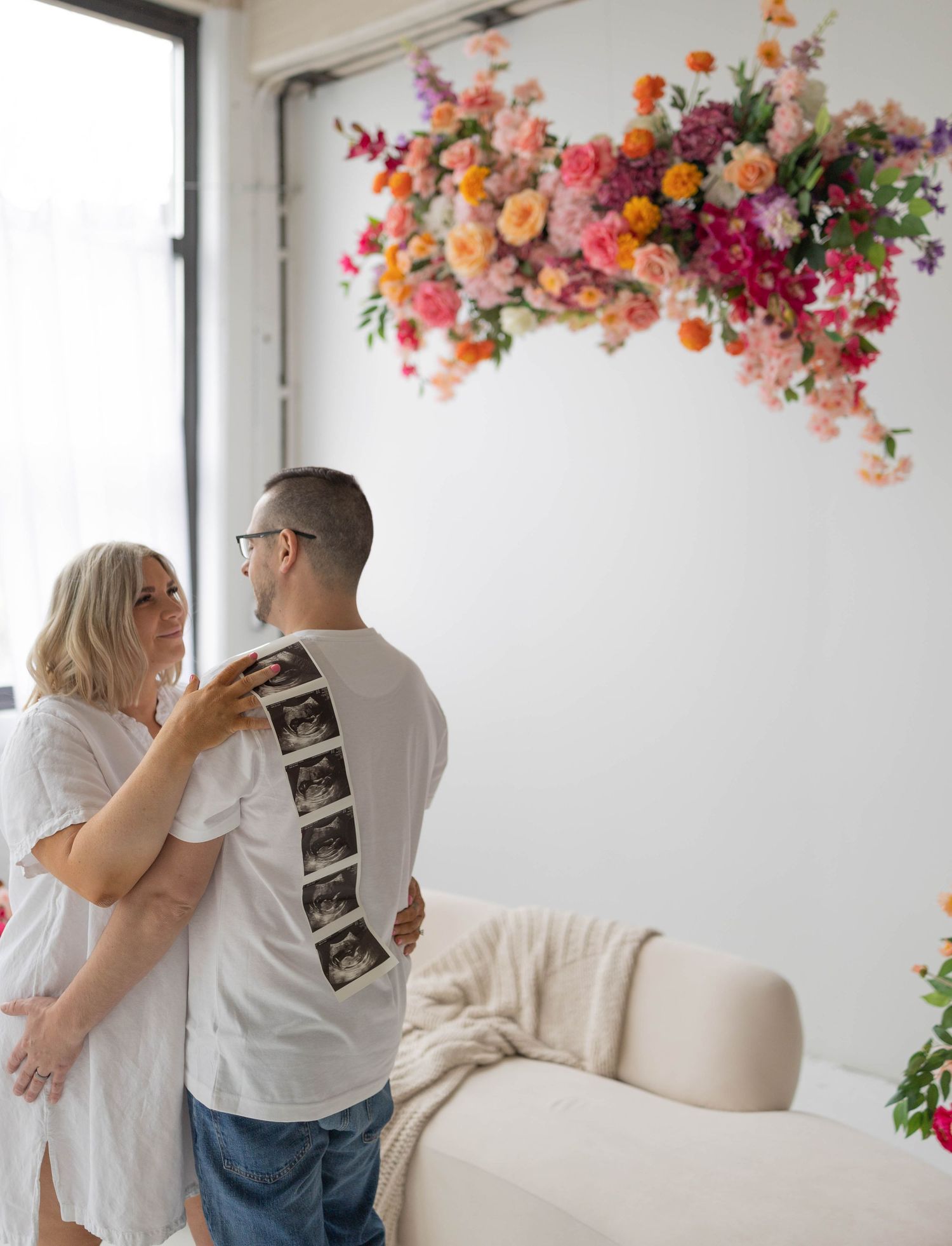 Expecting couple holds ultrasound photos under a colorful floral installation in a bright white room with cream furniture.