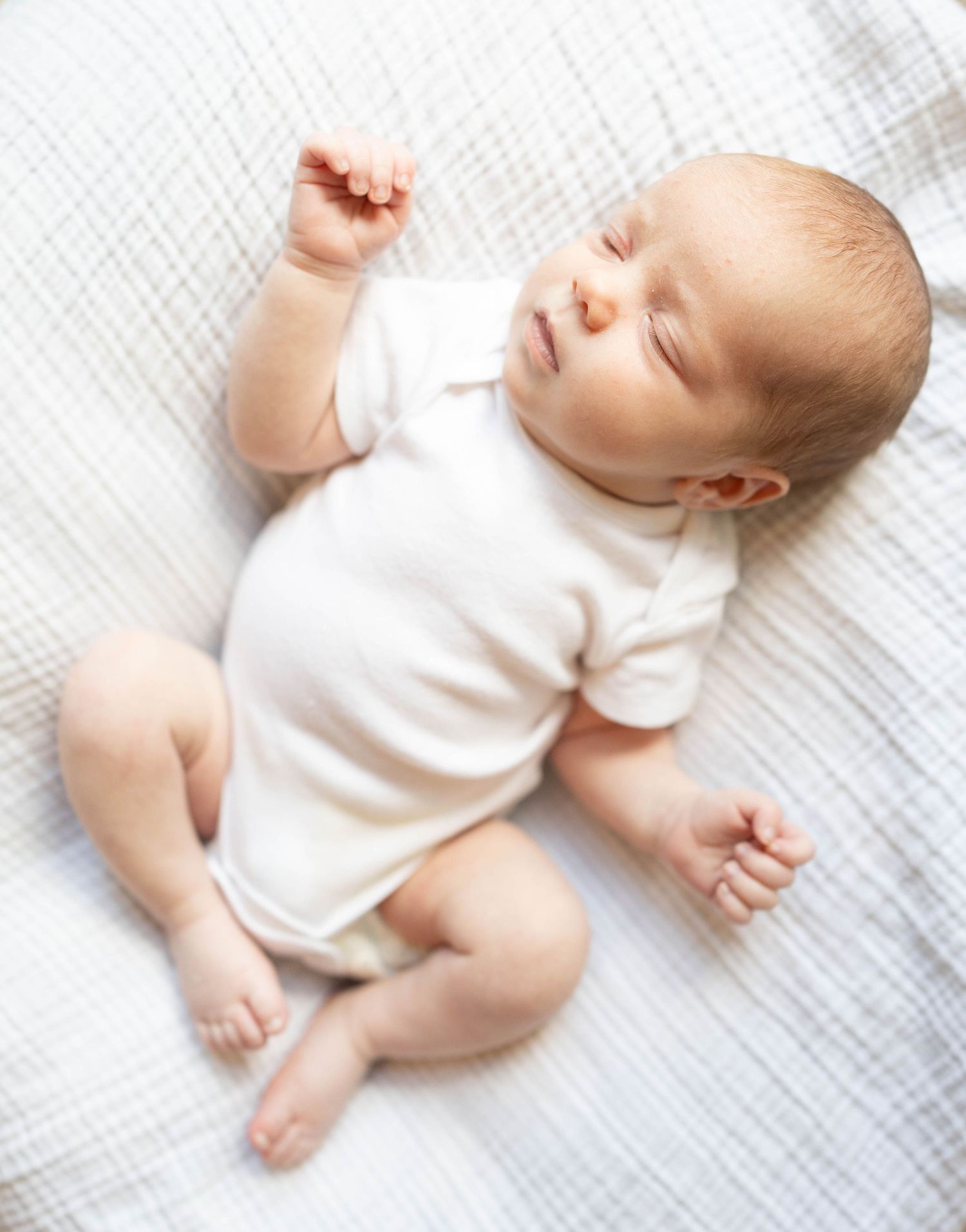 A sleeping baby wearing a white onesie lies peacefully on textured white bedding.