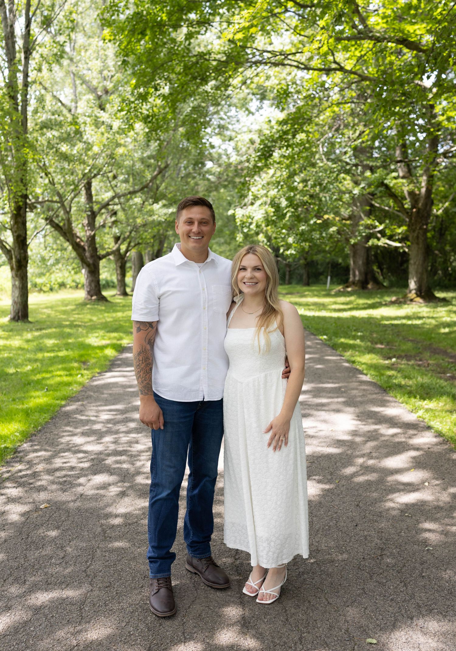Couple in white outfits smiling on a tree-lined path in a lush green park during summer.