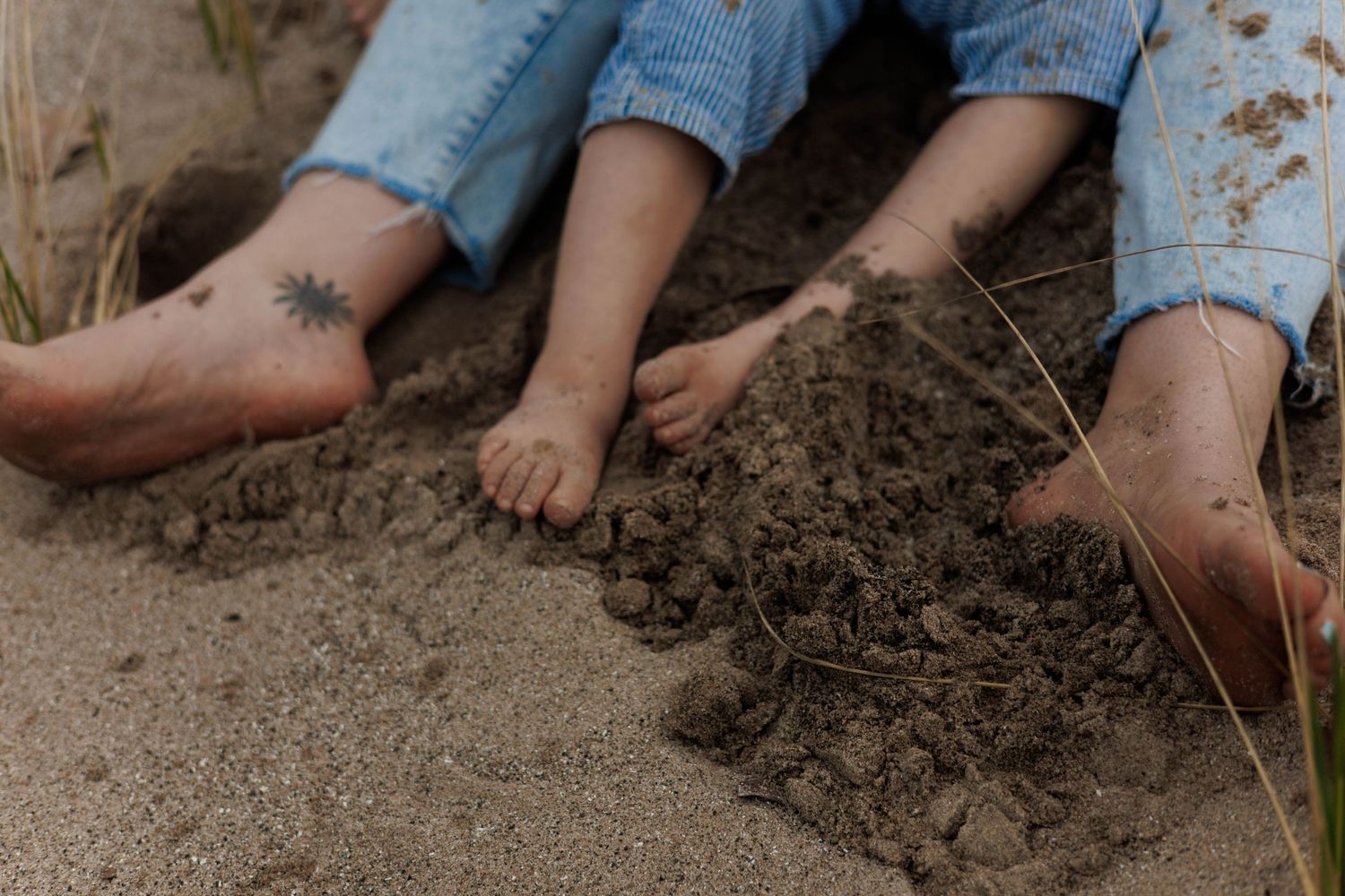 Bare feet of adults and a child playing in sandy soil outdoors, one ankle featuring a small floral tattoo.