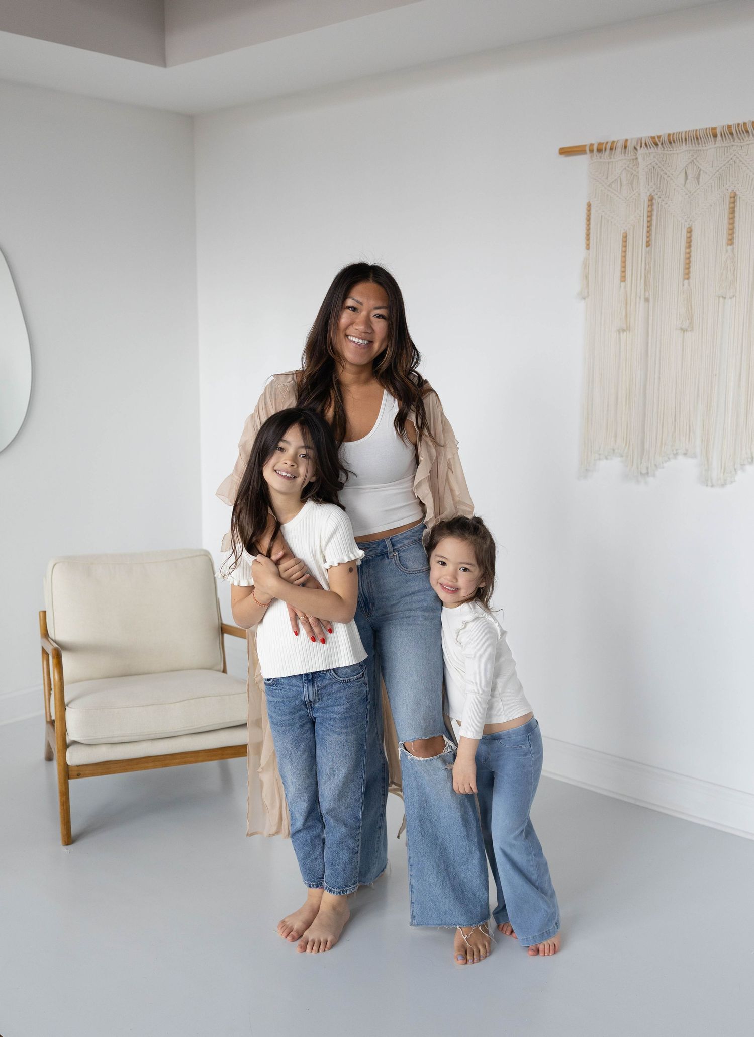 A woman and two young girls in matching white tops and jeans pose together in a bright, minimalist white room.