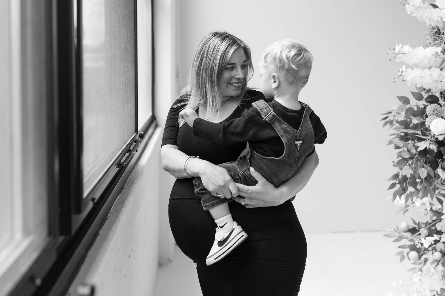 A person in black attire holds a small child while walking down a brightly lit hallway.