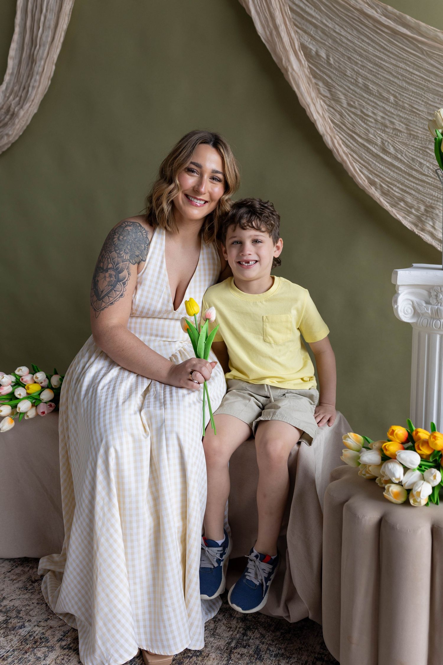 Smiling woman in checkered dress holding tulips poses with happy young boy in yellow shirt in floral studio.