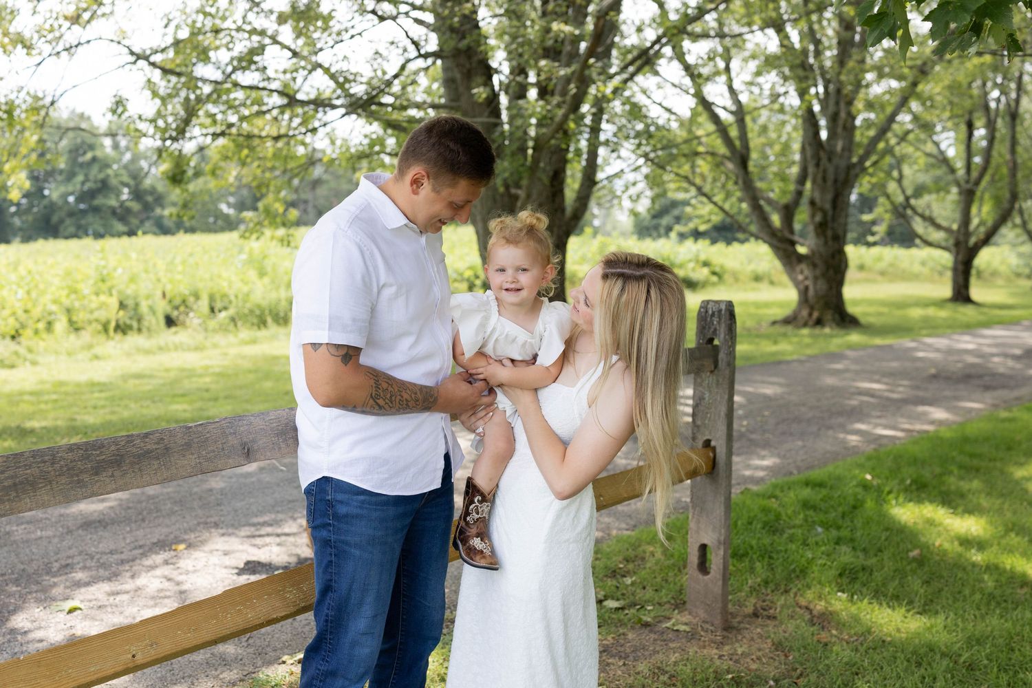 A young family with a toddler poses outdoors near a wooden fence surrounded by lush green trees on a sunny day.