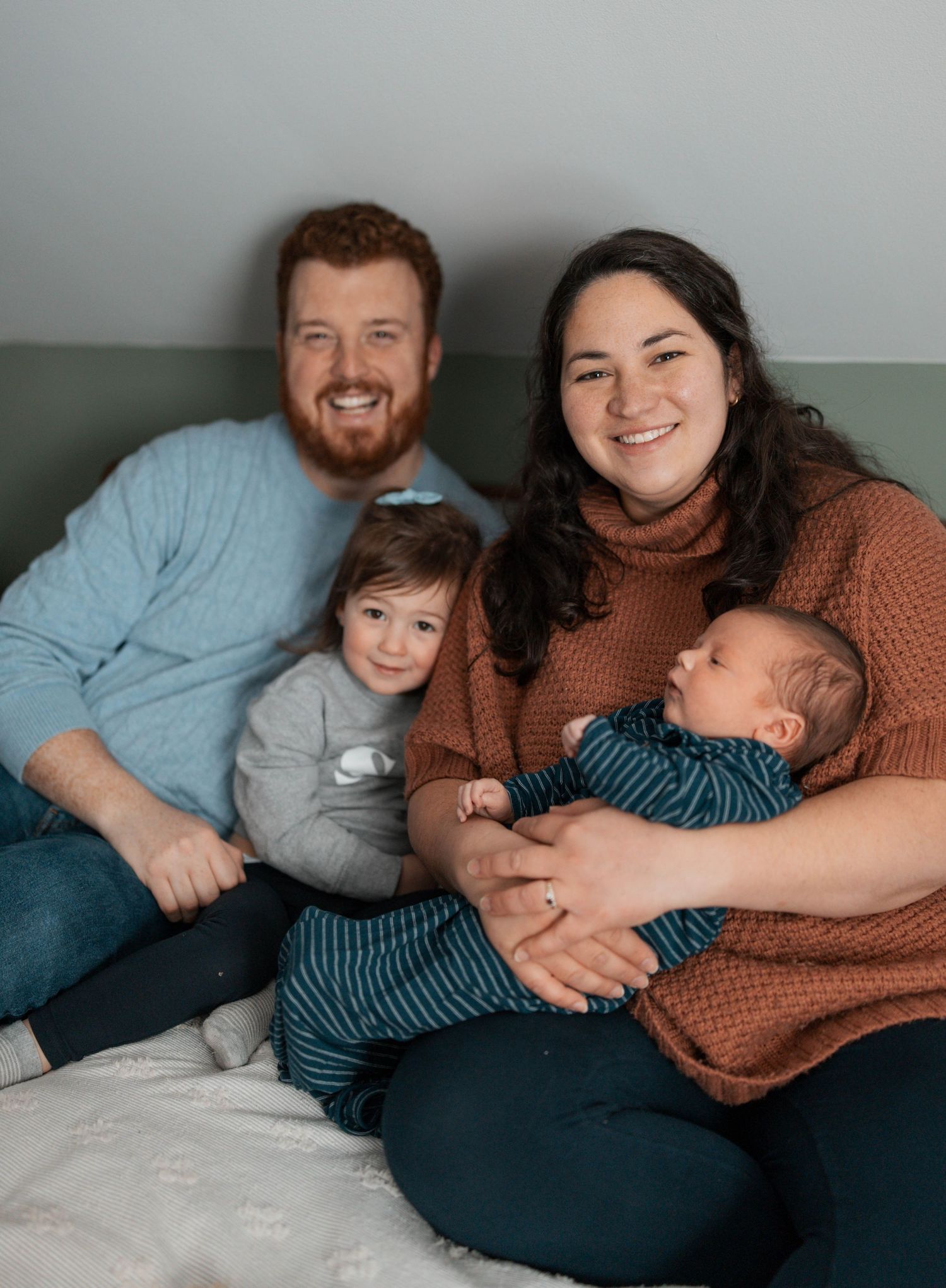 Family sits together on light gray couch with toddler and baby while wearing casual sweaters.