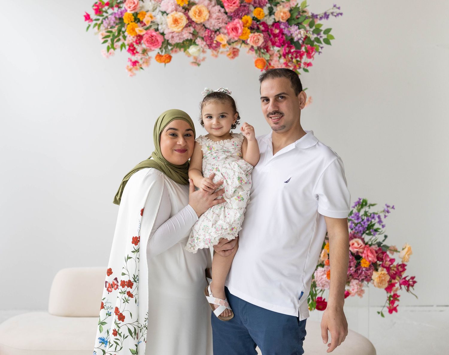A family poses under a vibrant floral arrangement with colorful pink and purple flowers overhead against a white backdrop.