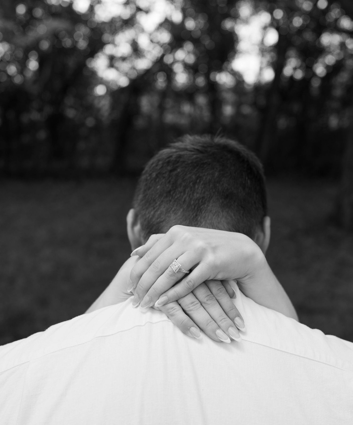 Black and white photo of a woman's hand with engagement ring resting on a man's neck from behind.