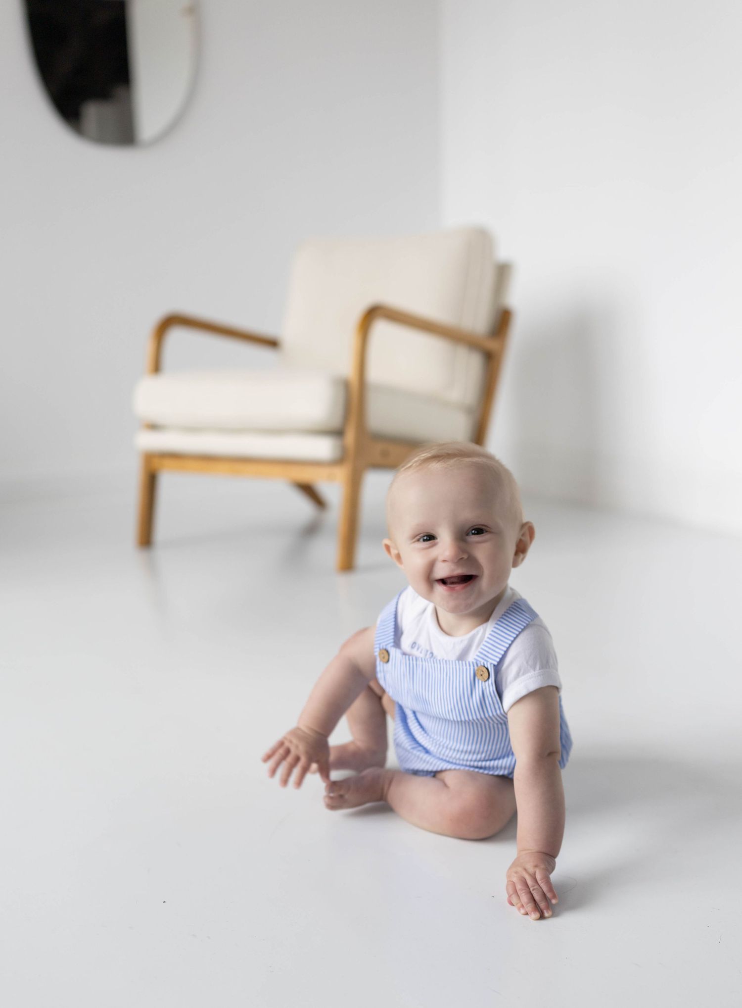 Smiling baby in blue overalls sitting on white floor with a wooden armchair in the background.