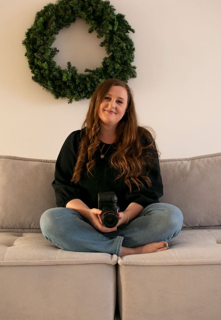 Person sitting cross-legged on a gray couch holding a camera with a green wreath on the wall above.