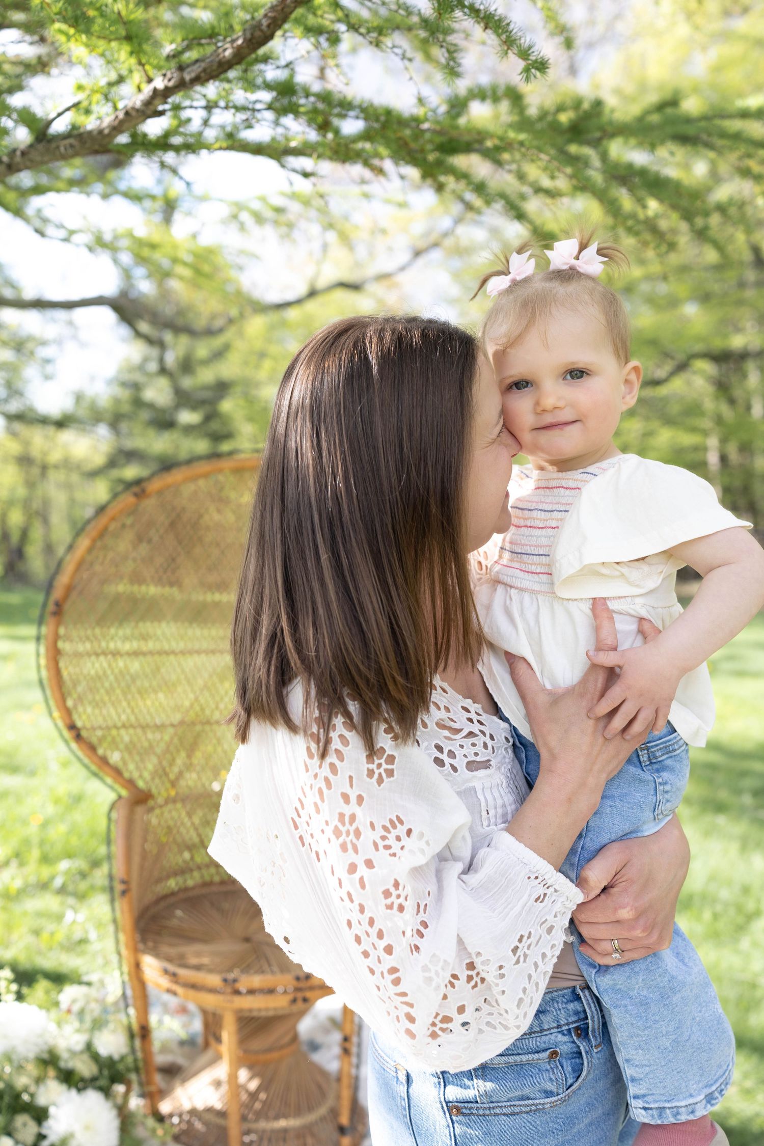 Woman in white lace top holds toddler in denim by wicker peacock chair in sunny garden setting.