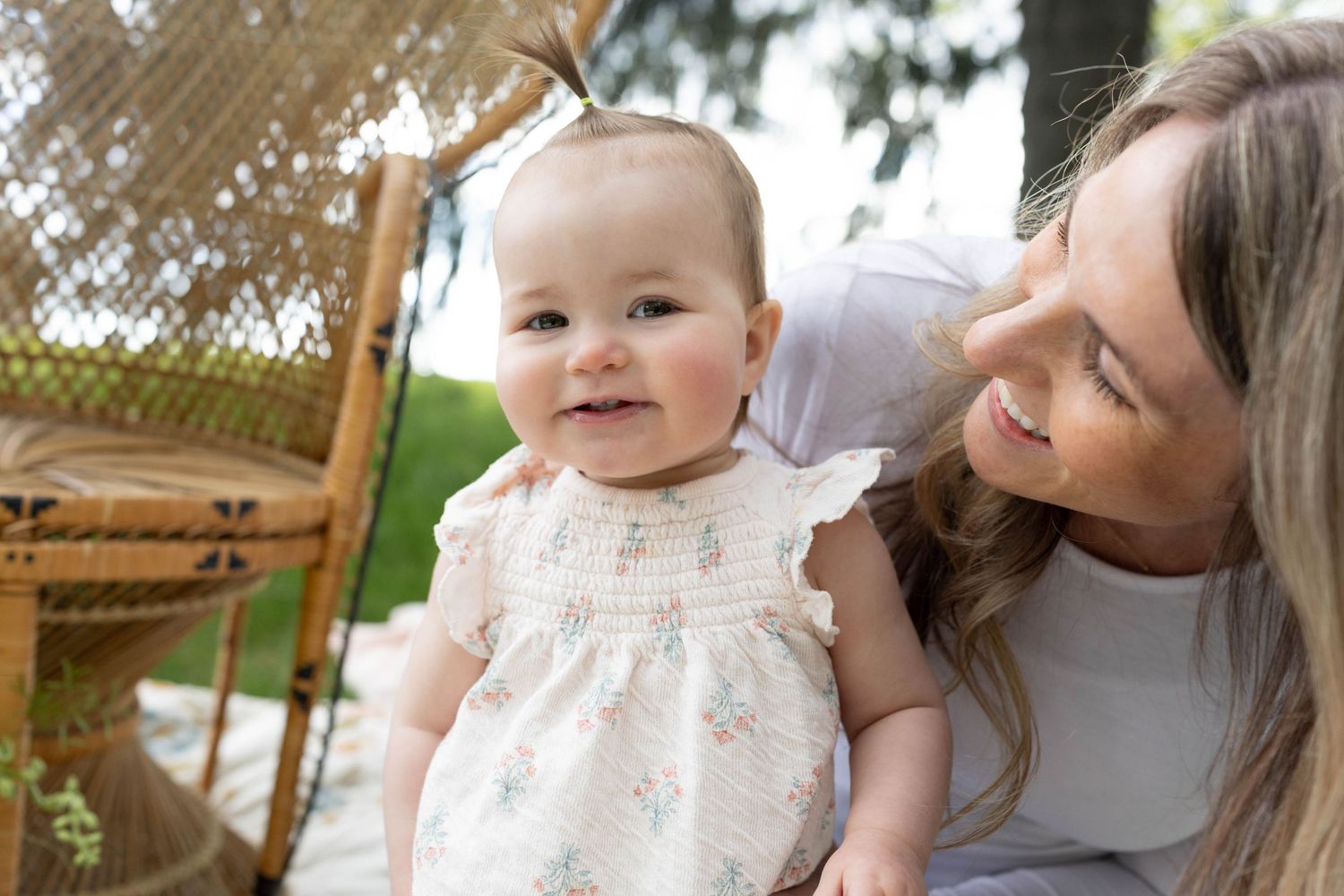 Happy infant in a white floral dress sits outdoors with a caregiver on a sunny day.