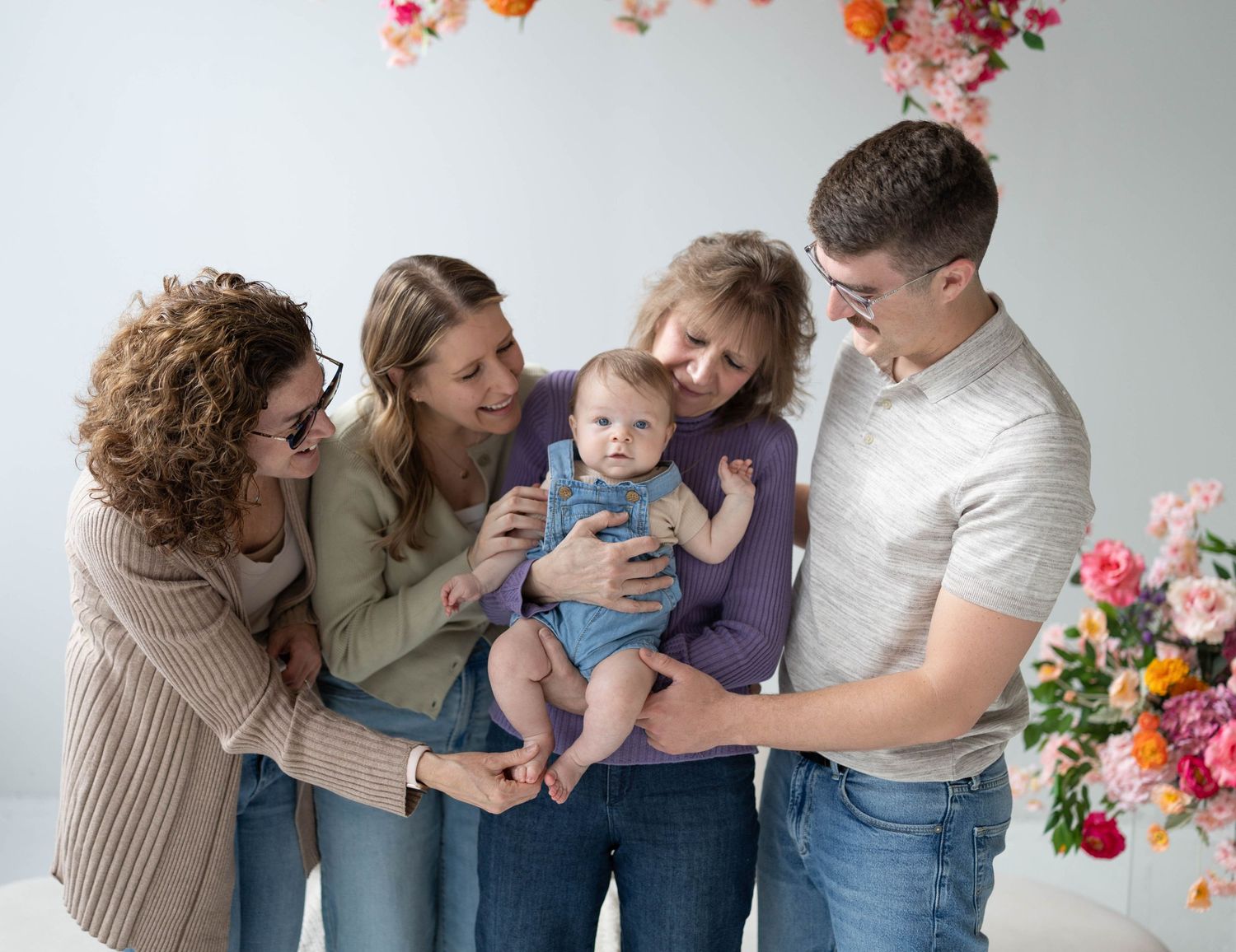 Three generations of family members in casual clothing enjoy a tender moment together against a backdrop of floral decorations.