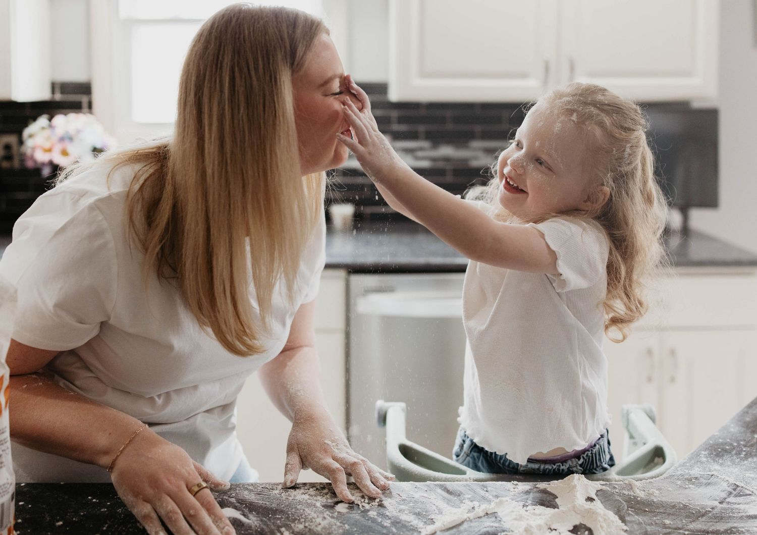 A child playfully touches someone's nose while they make a mess with flour in a bright kitchen setting.