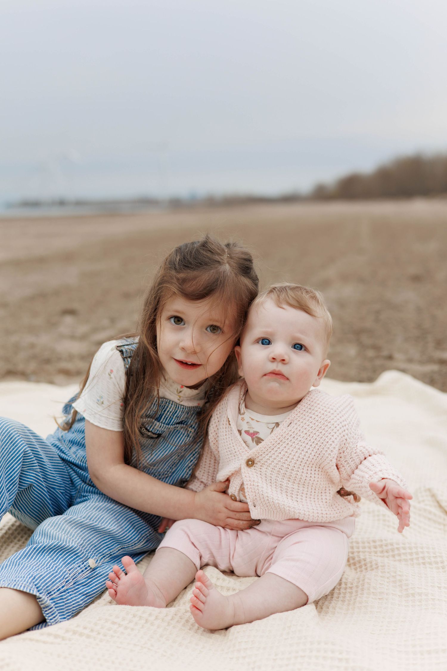 Two young sisters sitting on blanket on sandy beach, older girl hugging baby sibling.