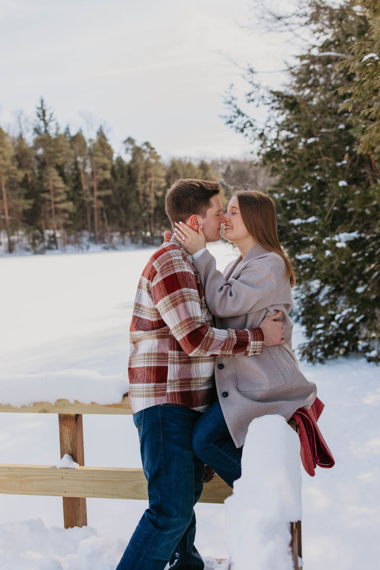 A couple embraces in a romantic winter setting with snow-covered trees and a wooden fence in the background.