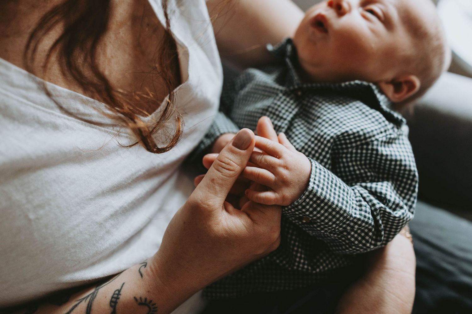 A tattooed mother tenderly holds her newborn baby's hand while cradling the infant in a cozy home setting.
