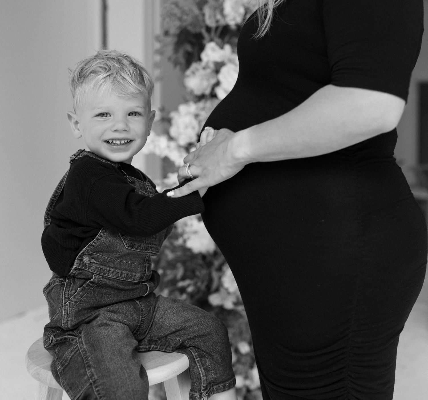 A young child in denim overalls holds a parent's hand while seated on a white stool with flowers in the background.