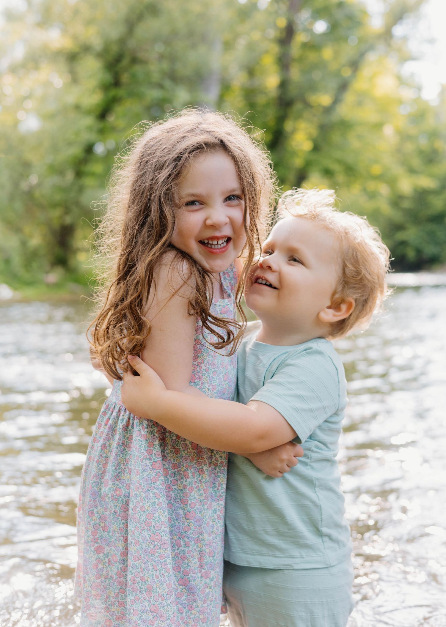 Two happy children hugging and laughing near a sunlit river, girl in floral dress and boy in mint shirt.