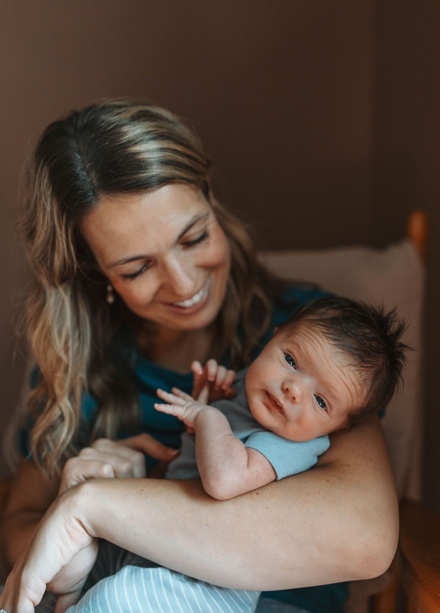Smiling mother holding her newborn baby in a blue onesie, captured in a warm indoor setting.