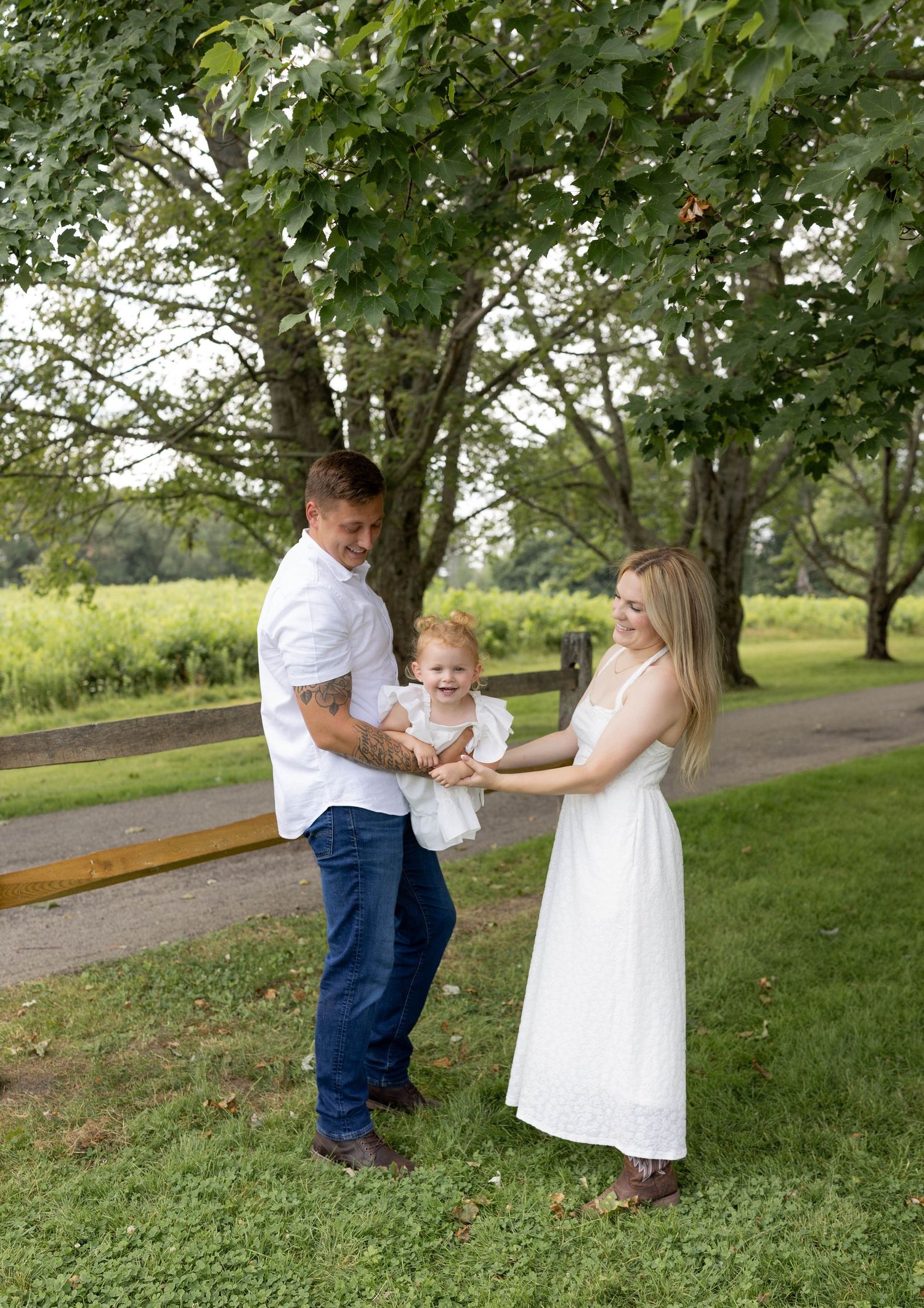 A happy family of three, with parents in white outfits and jeans, playfully holding their toddler outdoors near lush green trees.