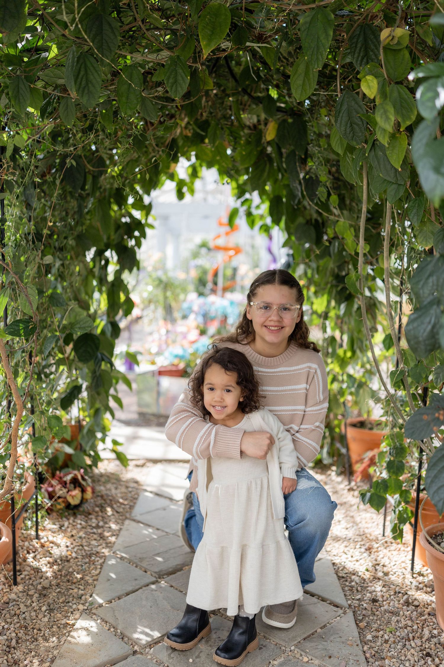 Two people pose together beneath a leafy garden archway on a stone path with potted plants in the background.