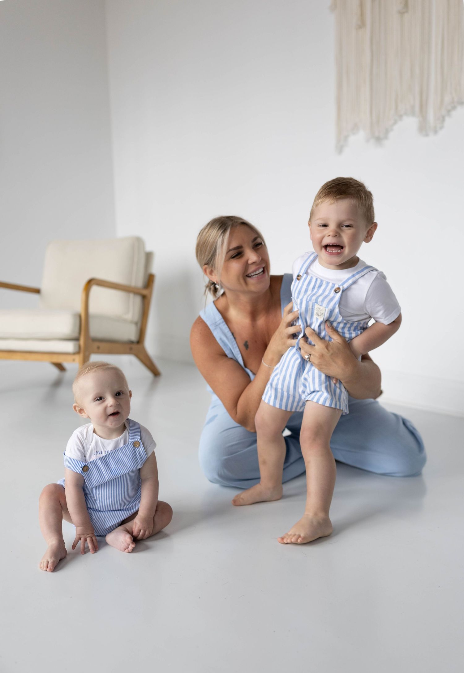 A smiling woman kneels on a white floor holding a laughing toddler, with a baby sitting nearby, all wearing matching blue outfits.