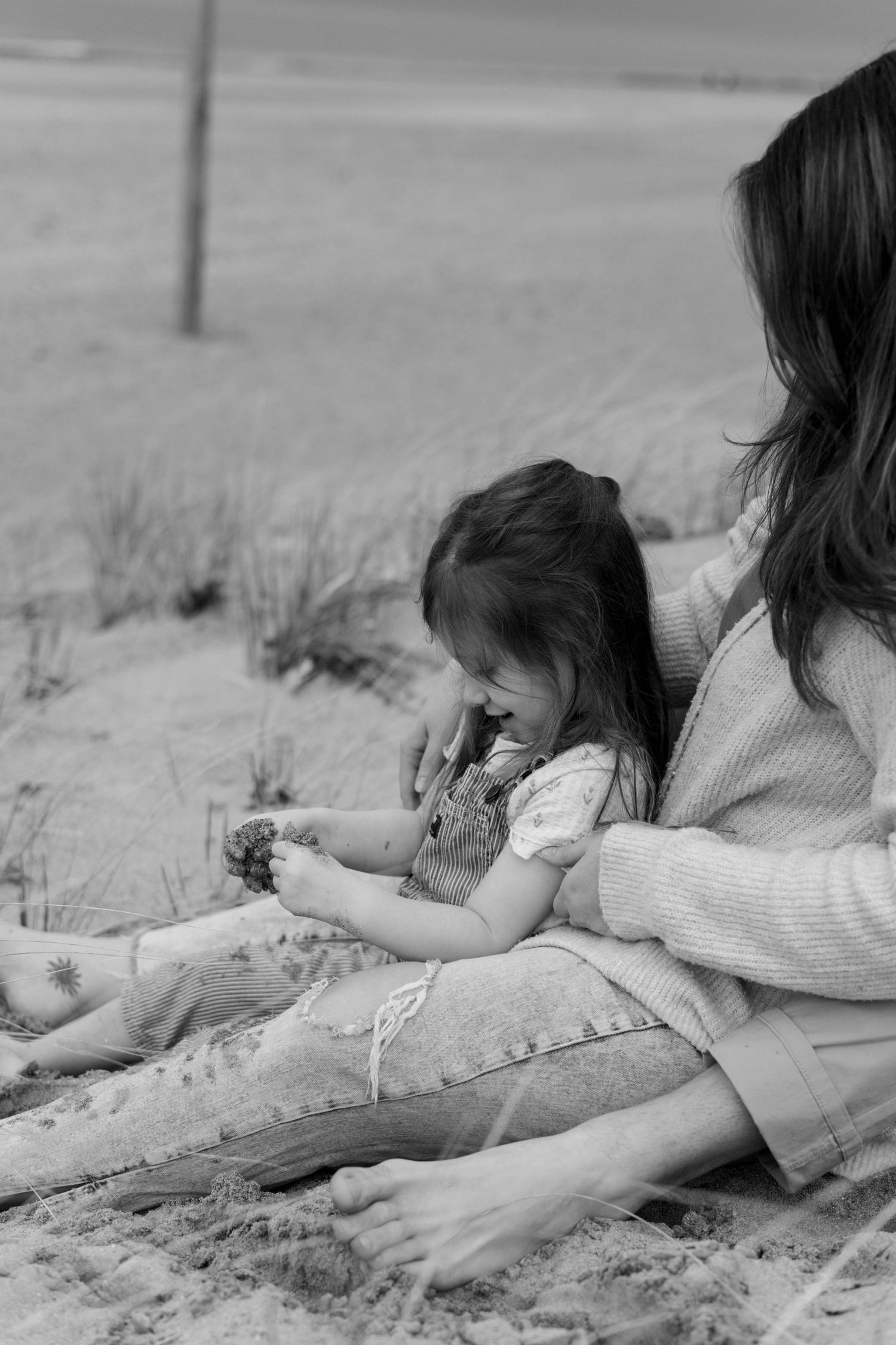 Black and white photo of a mother and young daughter sitting on sandy beach, child in overalls examining something in her hands.