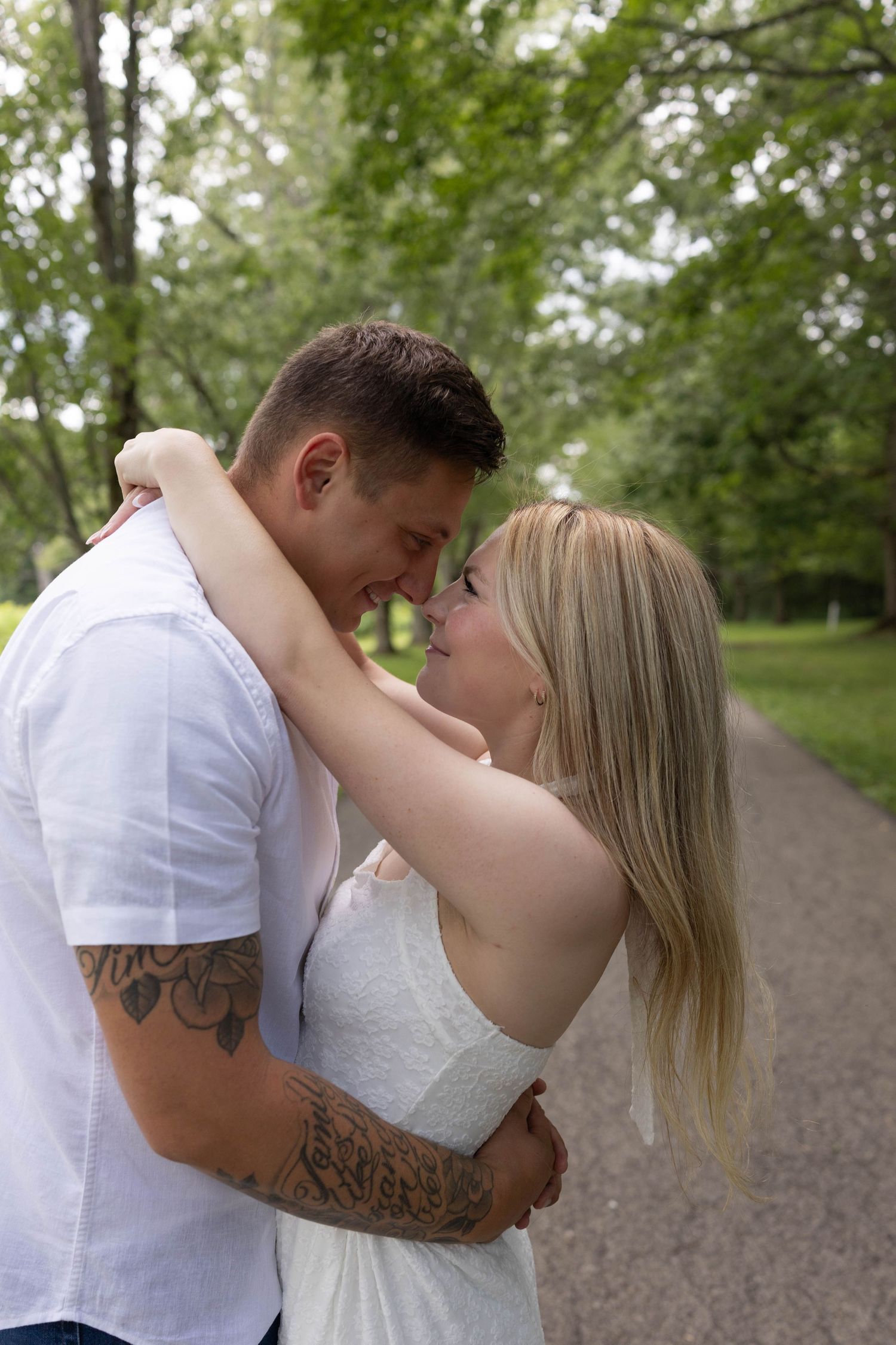 Romantic couple embracing and touching foreheads on a scenic park pathway surrounded by green trees.