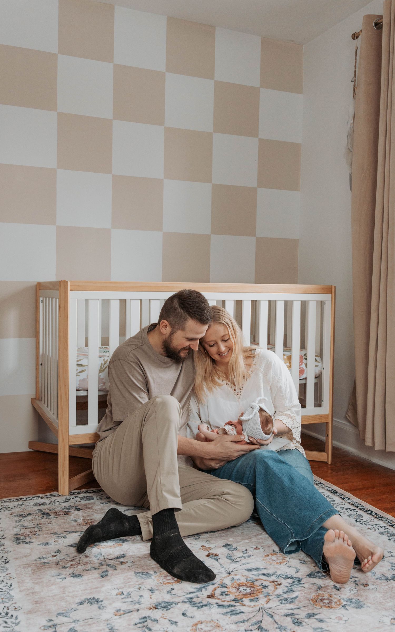 A couple sits on a patterned rug near a wooden crib in a nursery with checkered walls.