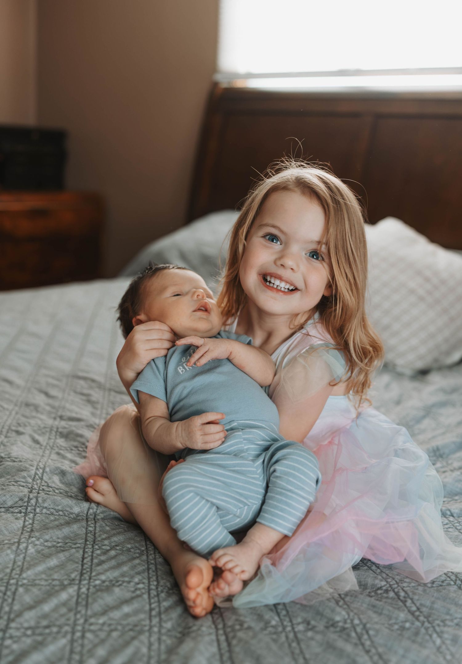 Smiling young girl in a pink tutu sitting on a bed while lovingly holding her newborn baby sibling.