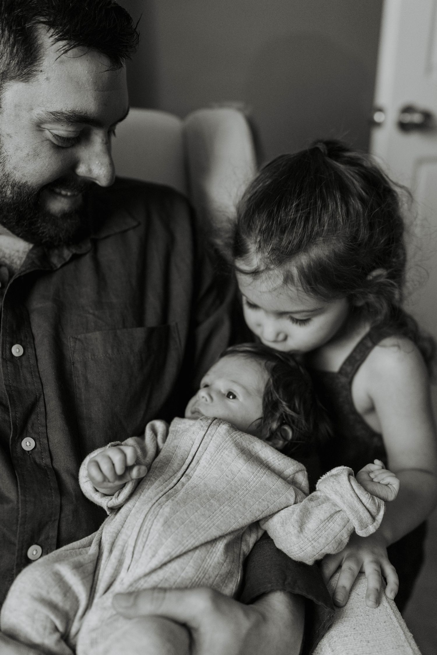 Bearded father smiles at newborn baby while young toddler sibling leans in to give a gentle kiss, black and white photo.