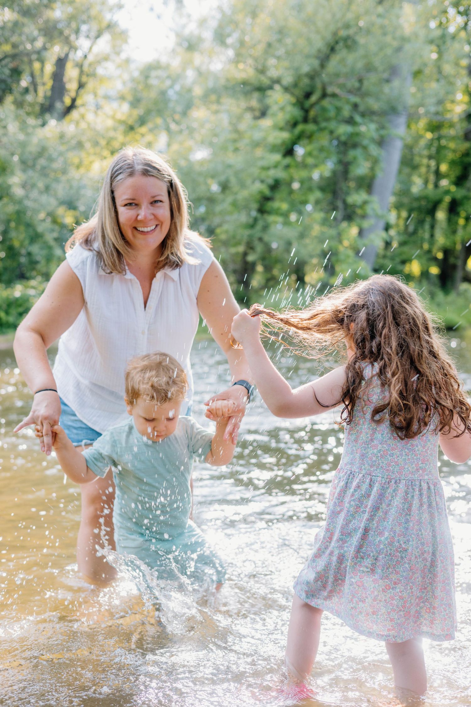A smiling mother plays with her two young children splashing in a shallow river surrounded by lush green trees.