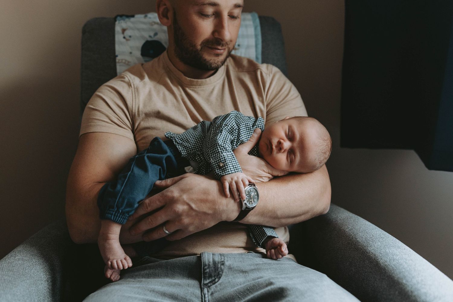 A bearded father in a beige shirt gently holds his sleeping newborn baby while sitting in a gray armchair.