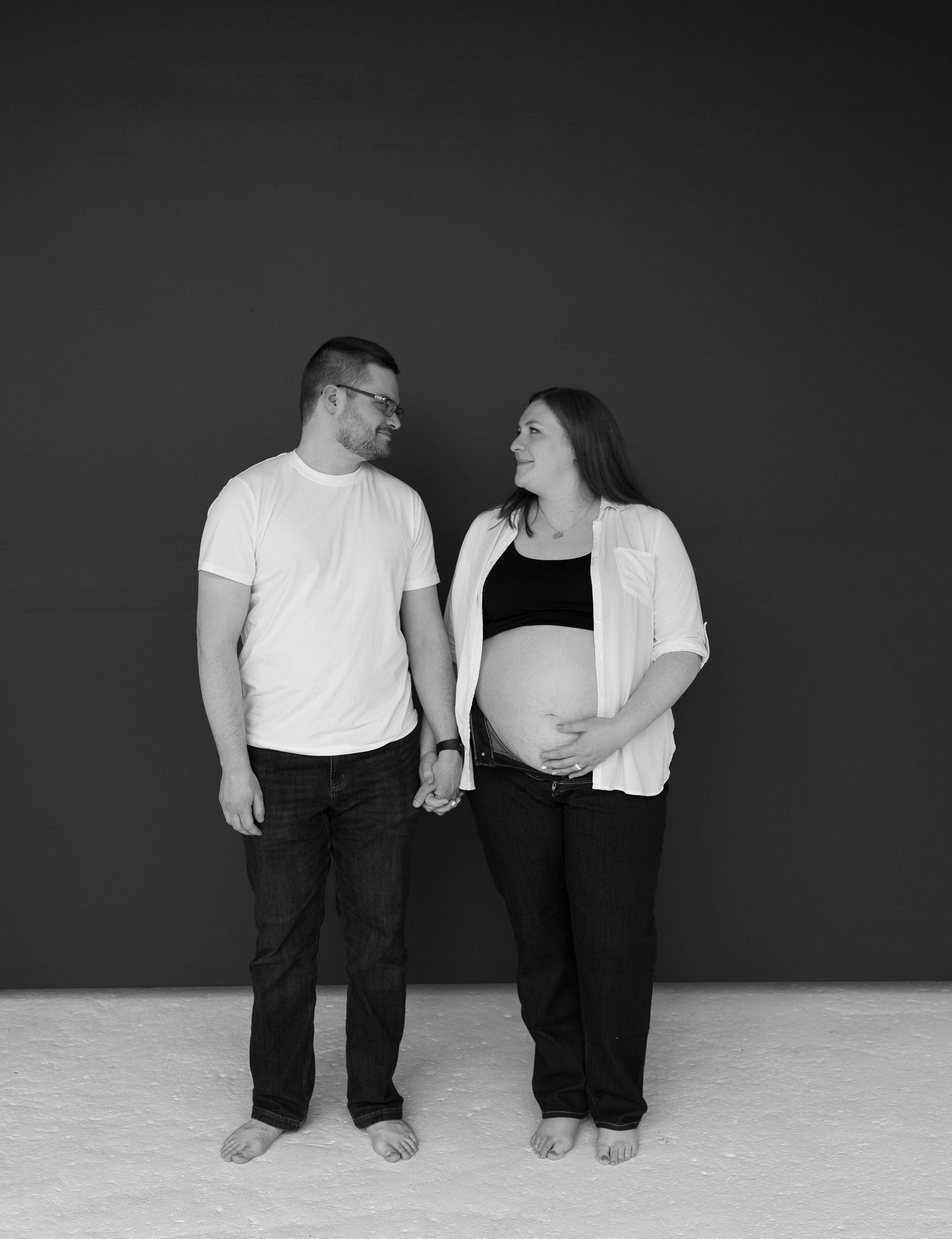 A black and white studio maternity photo features two people holding hands and smiling at each other against a dark backdrop.