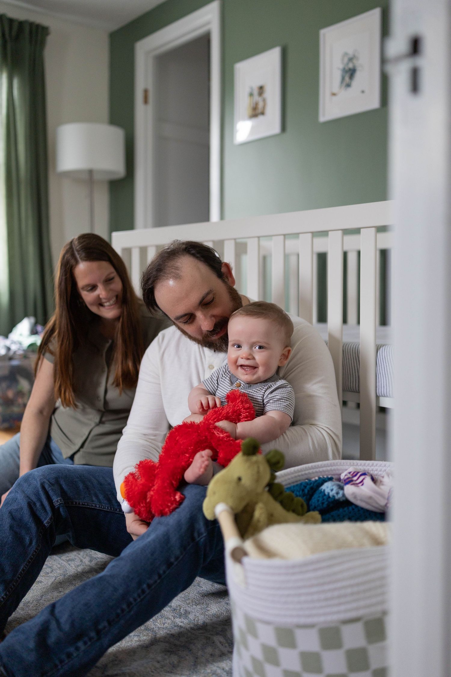 A family sits on a bed with toy stuffed animals while a baby plays with a red plush toy in a cozy bedroom.