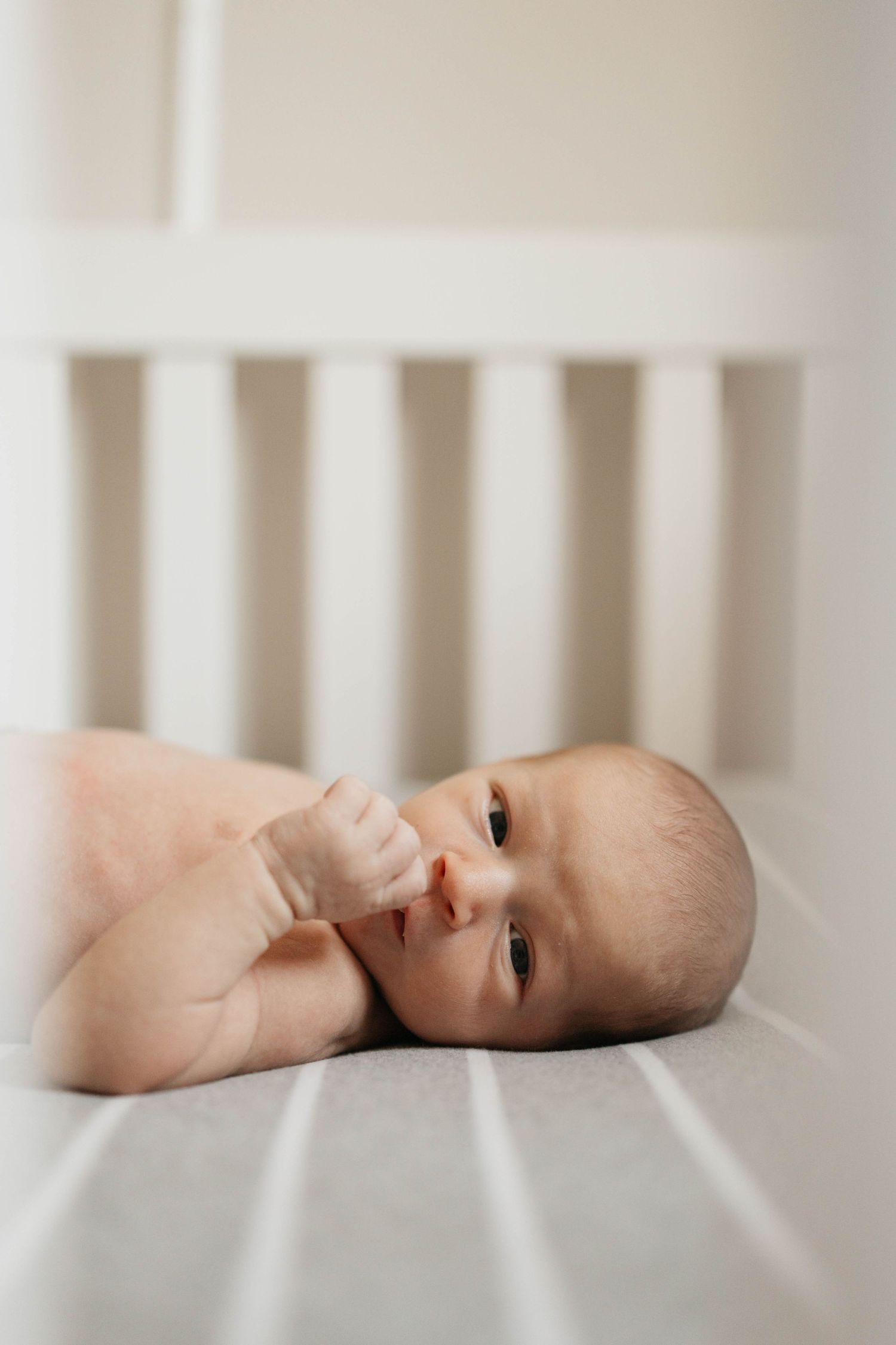 A baby lies on their side in a white crib, looking toward the camera with a gentle expression.