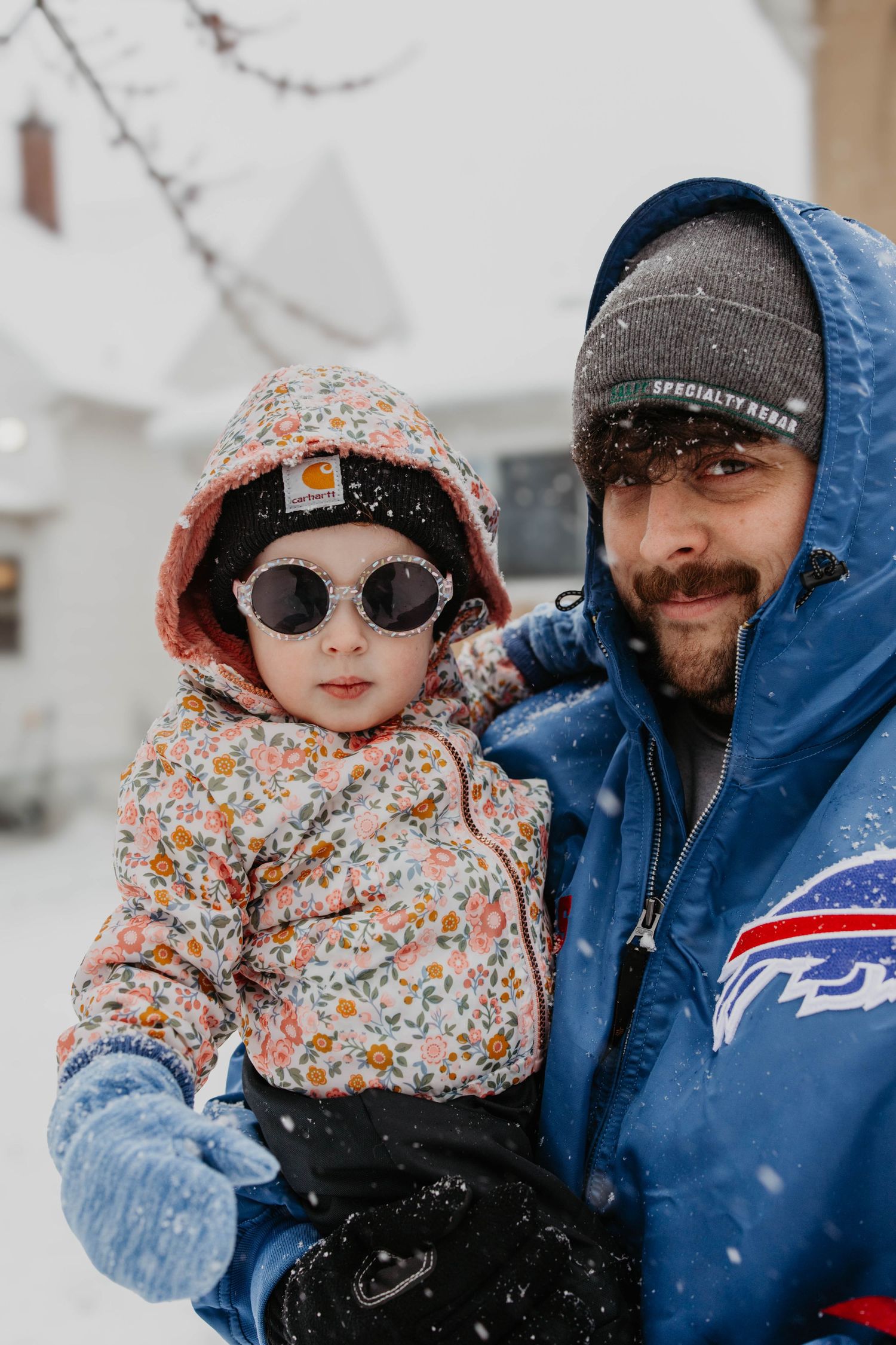 A parent and child bundled up in winter coats and hats stand together as snow falls around them.