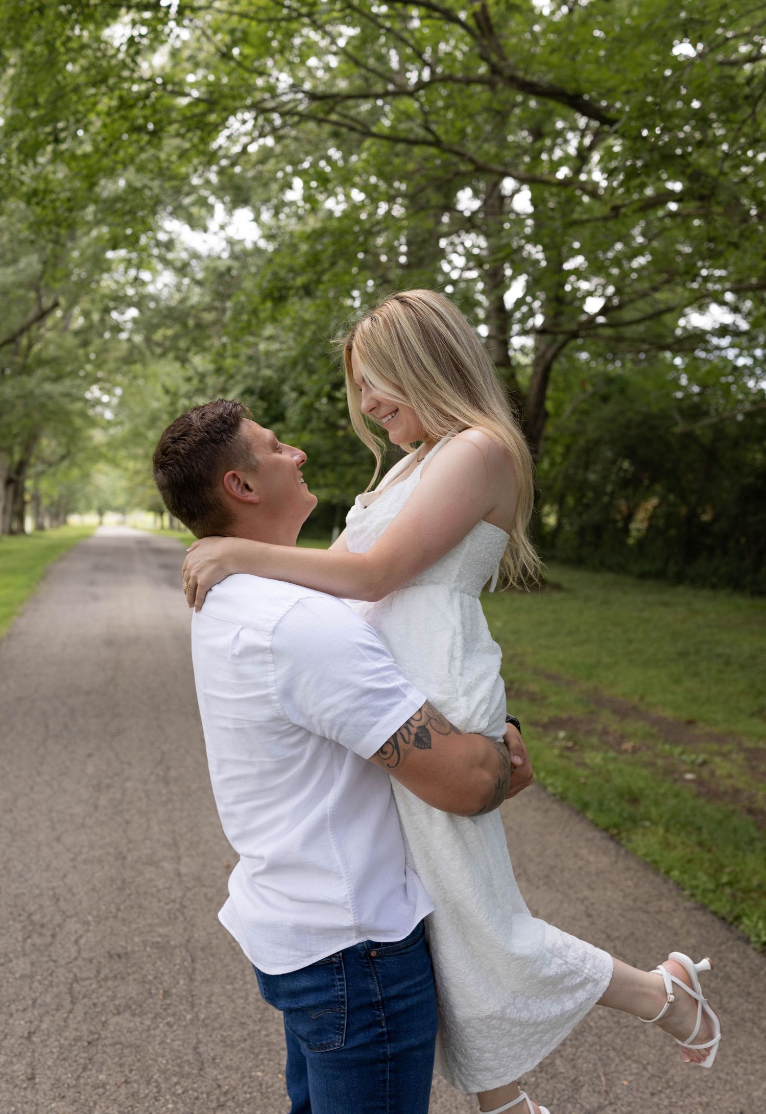 A couple in white outfits sharing a joyful moment on a tree-lined path, as the man lifts the woman.
