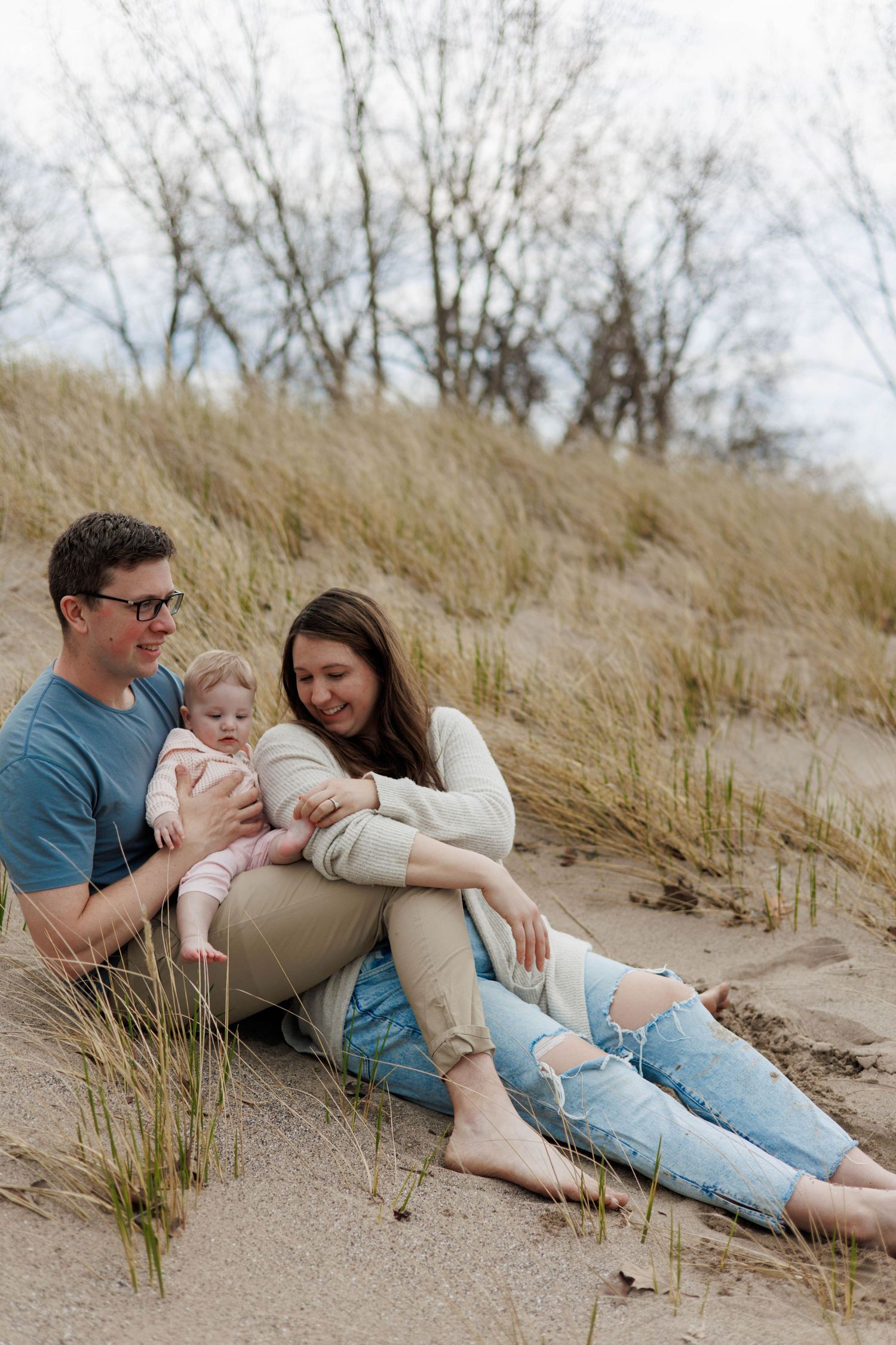 A young family sits on sandy beach dunes, parents smiling at their baby, surrounded by tall grass and bare trees.