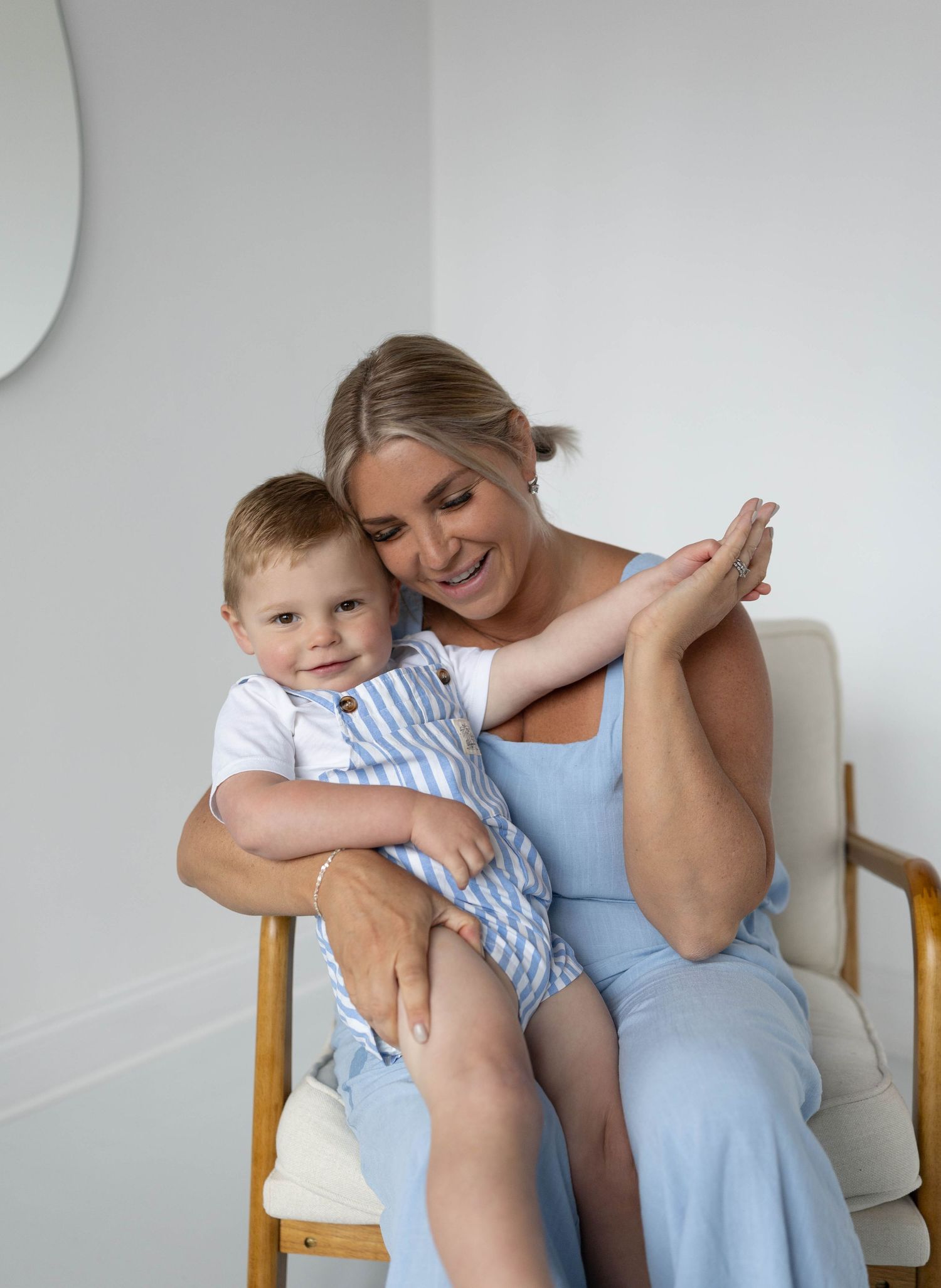Smiling blonde woman in blue dress sitting on chair holding a happy toddler boy in striped romper.