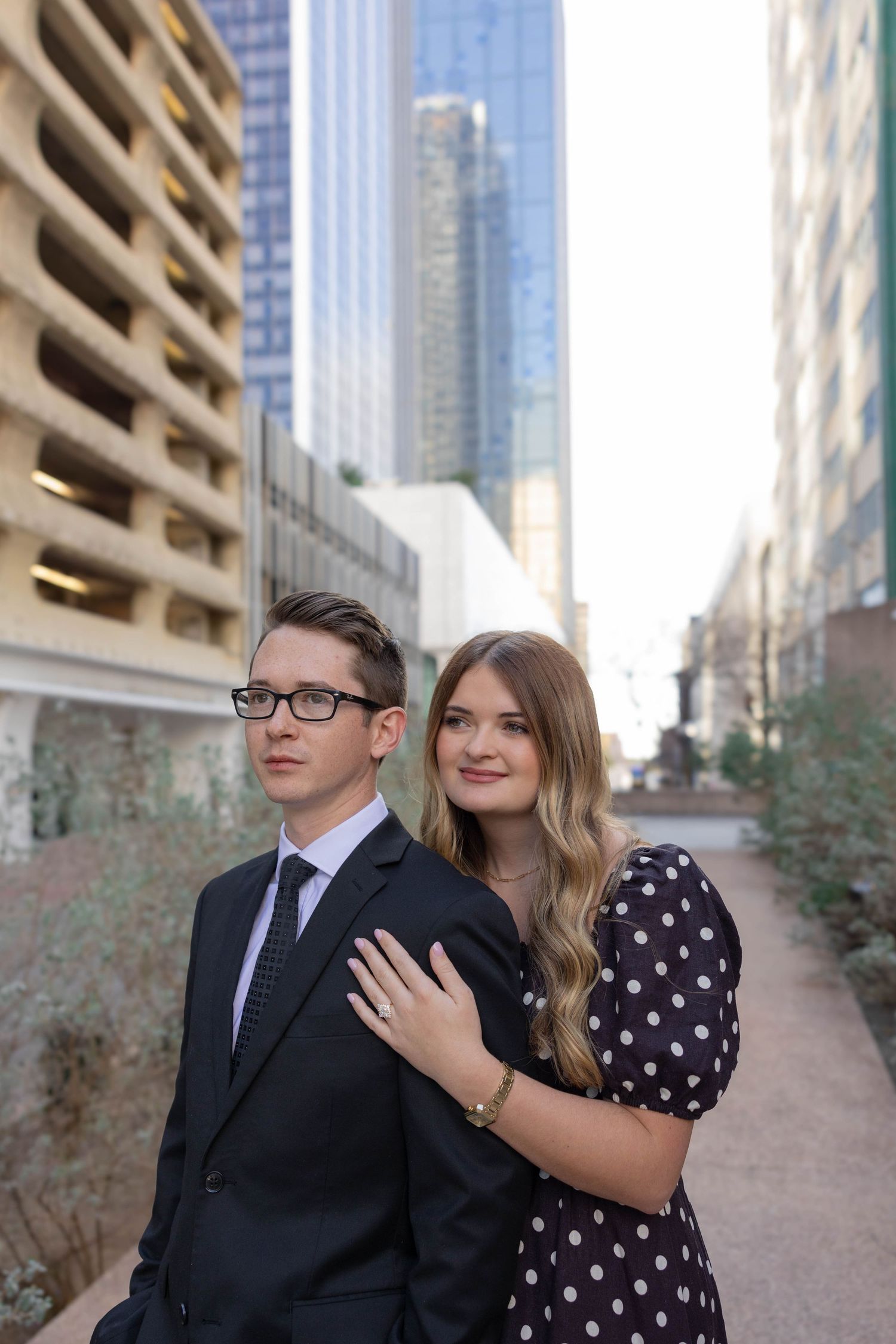 A couple in formal attire poses together on an urban street with skyscrapers and a parking garage in the background.