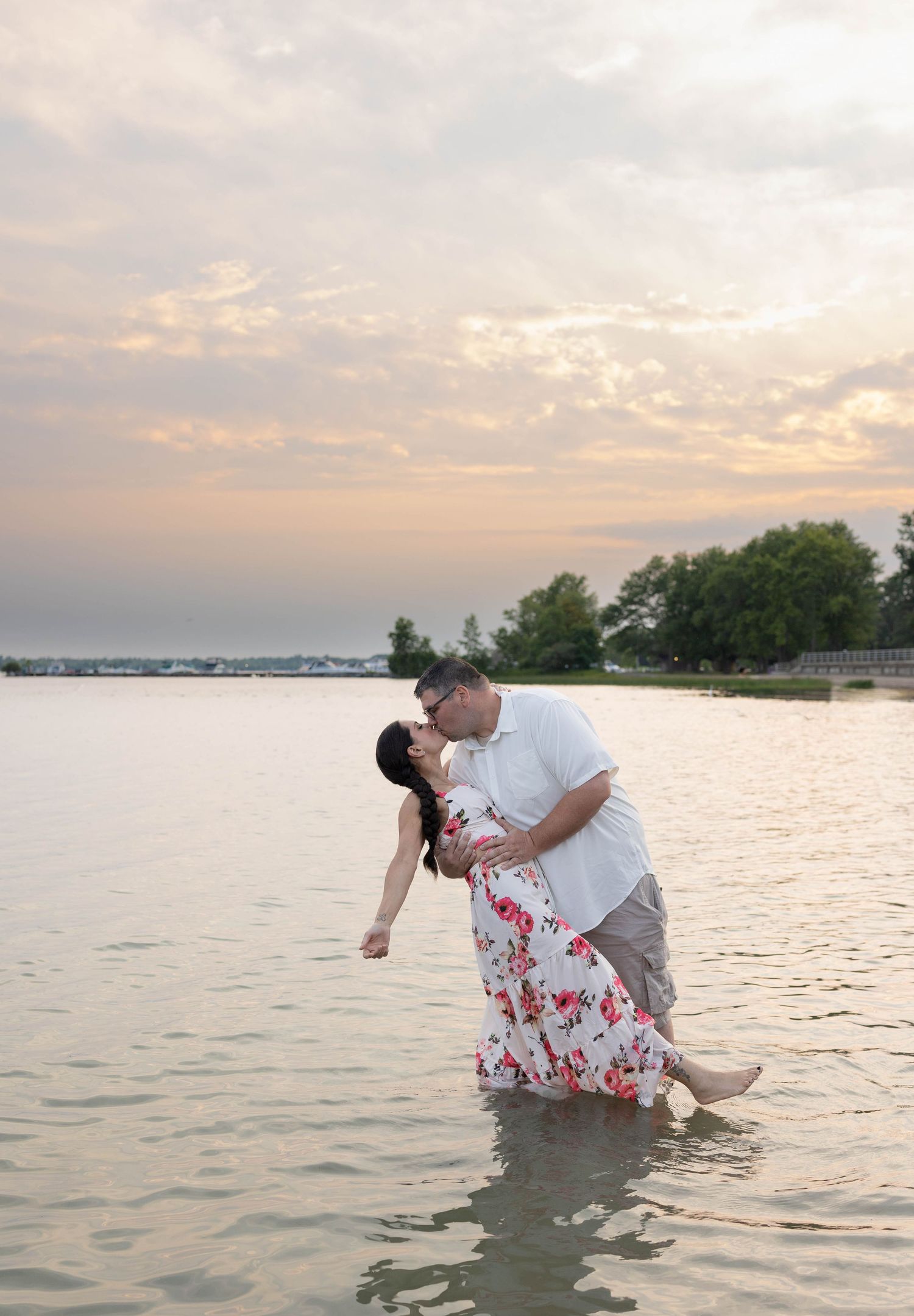 Couple sharing a romantic kiss while standing in shallow water at sunset, woman in floral maxi dress, man in white shirt.