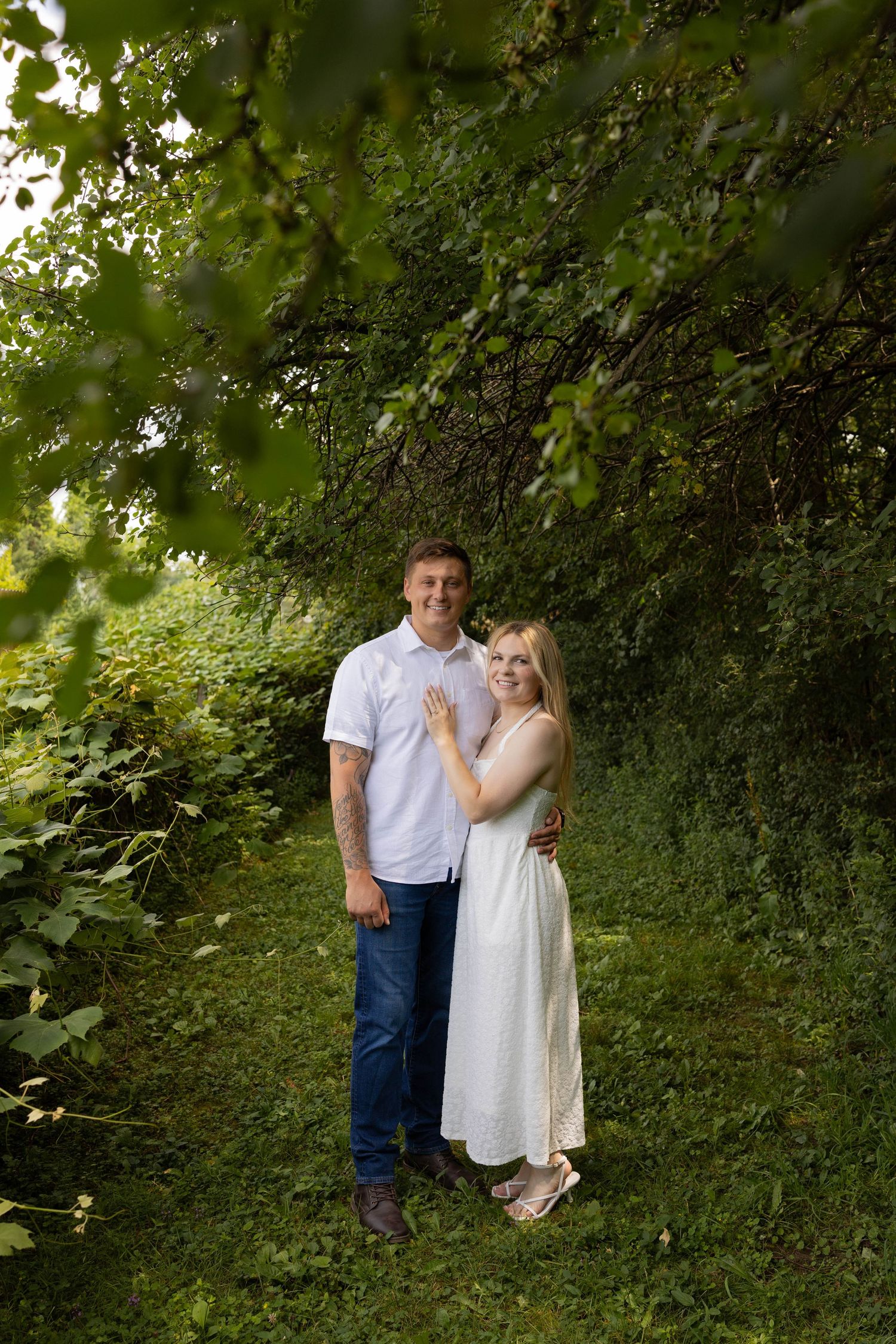 A smiling couple poses together in a lush green garden, woman in white dress, man in jeans and white shirt.