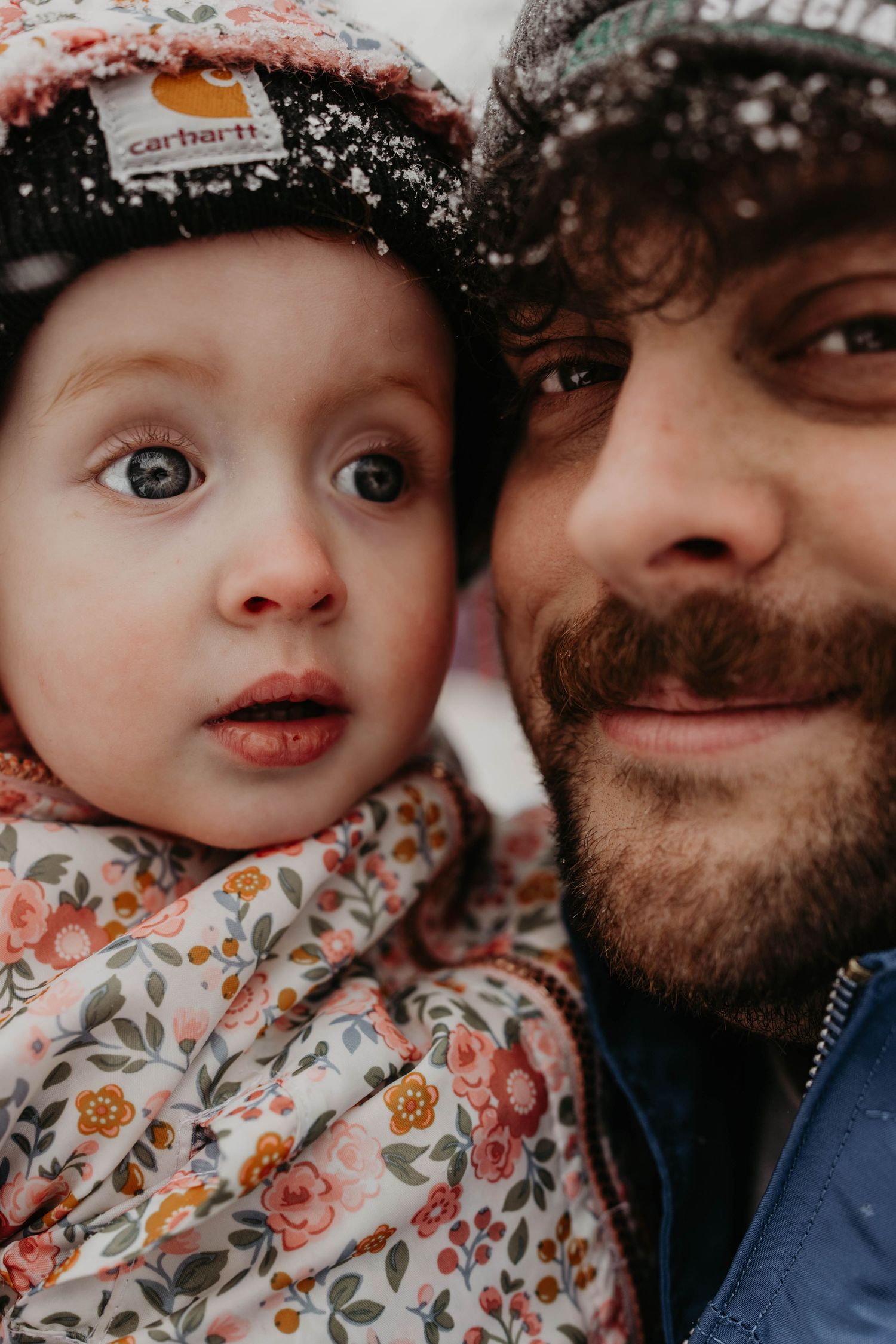 A close-up winter portrait showing a bearded person and small child wearing knit hats in the snow.