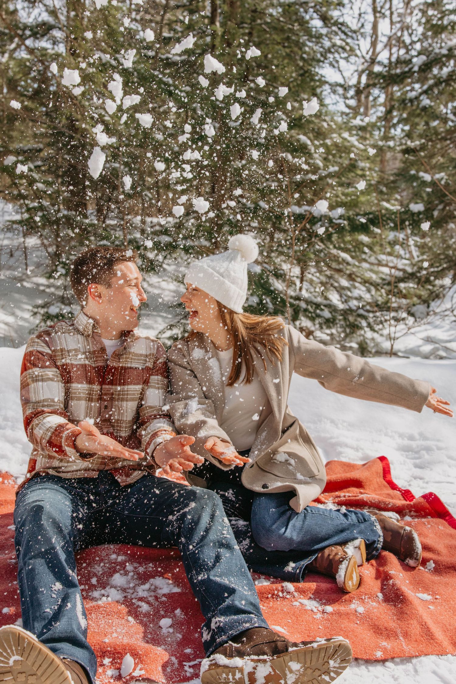 Two people sitting on an orange blanket in the snow while snowflakes fall around evergreen trees in winter.