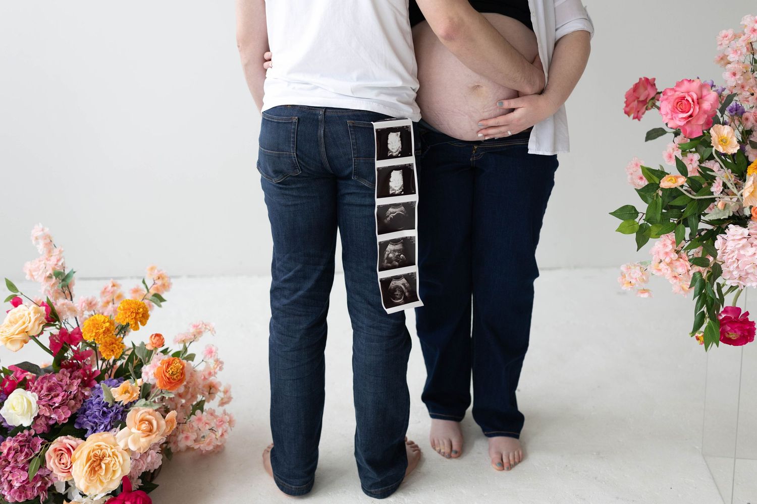 Couple stands close together against white wall with colorful floral arrangement featuring pink and orange blooms.