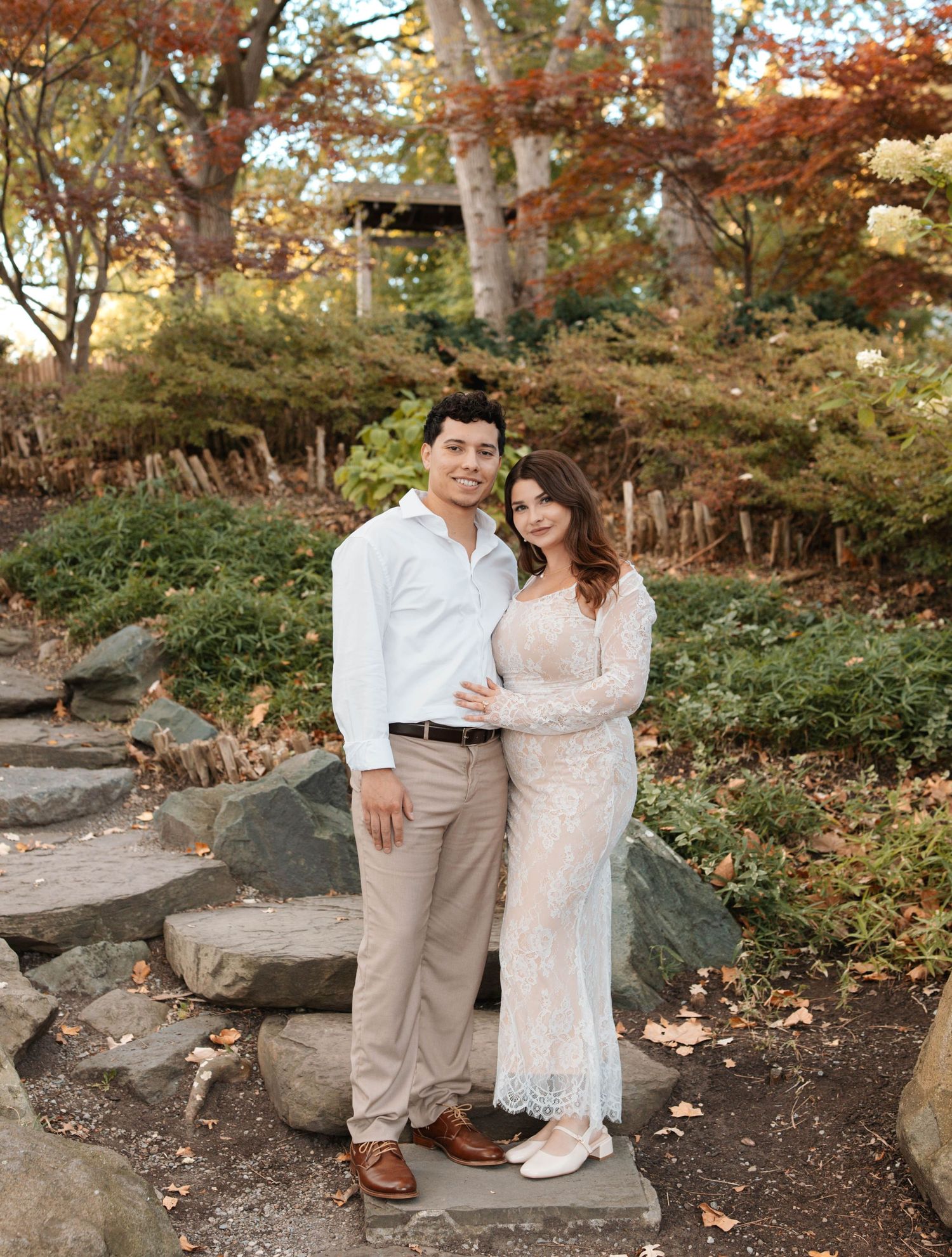 A couple sits together on stone steps surrounded by autumn foliage in a romantic outdoor setting.