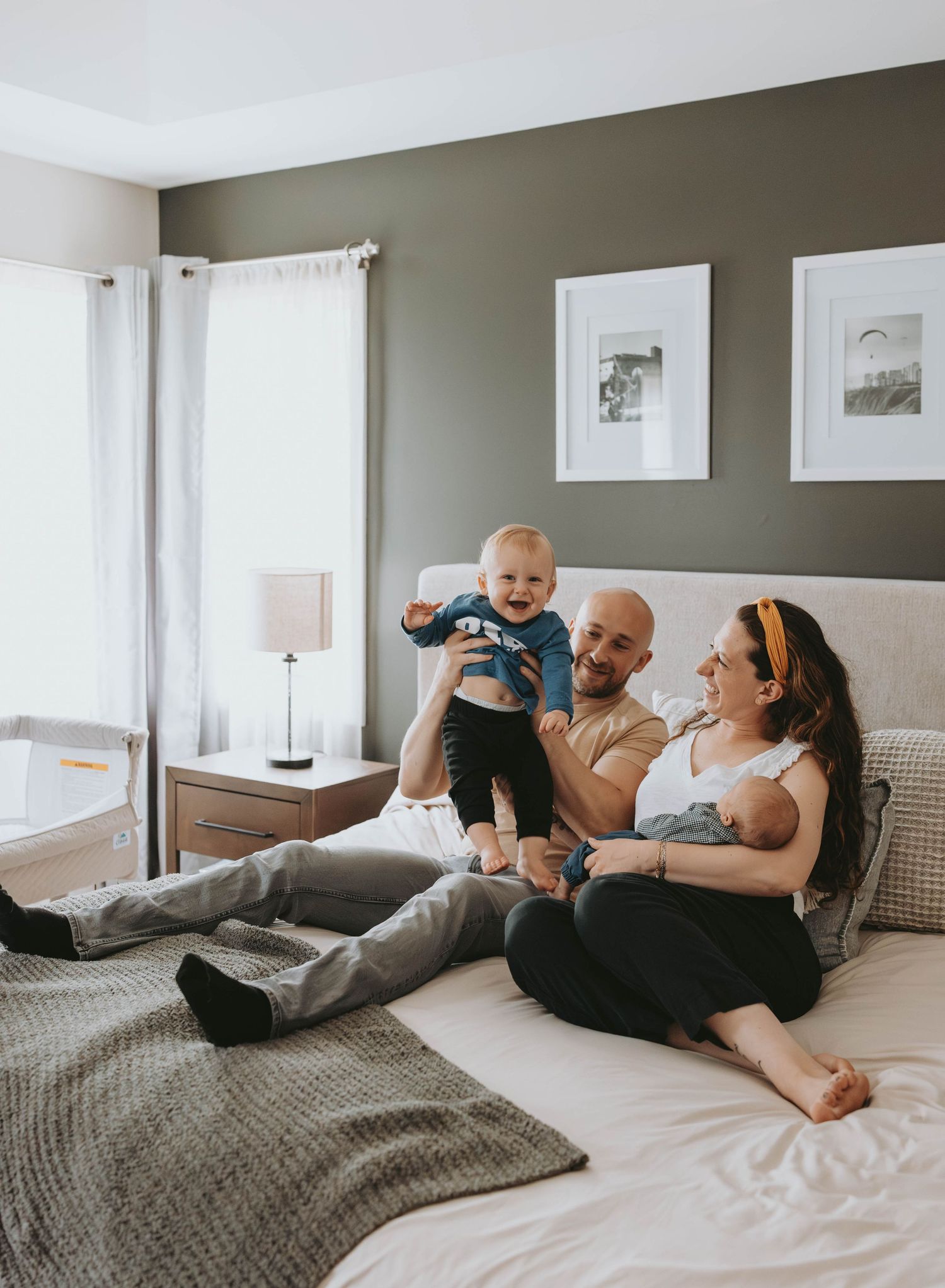 Happy family with bald dad, mom in headband, and laughing baby sitting on bed in cozy gray bedroom.