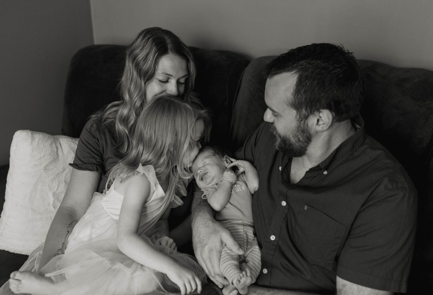 Black and white family photo of parents with toddler girl and newborn baby sitting on a couch.