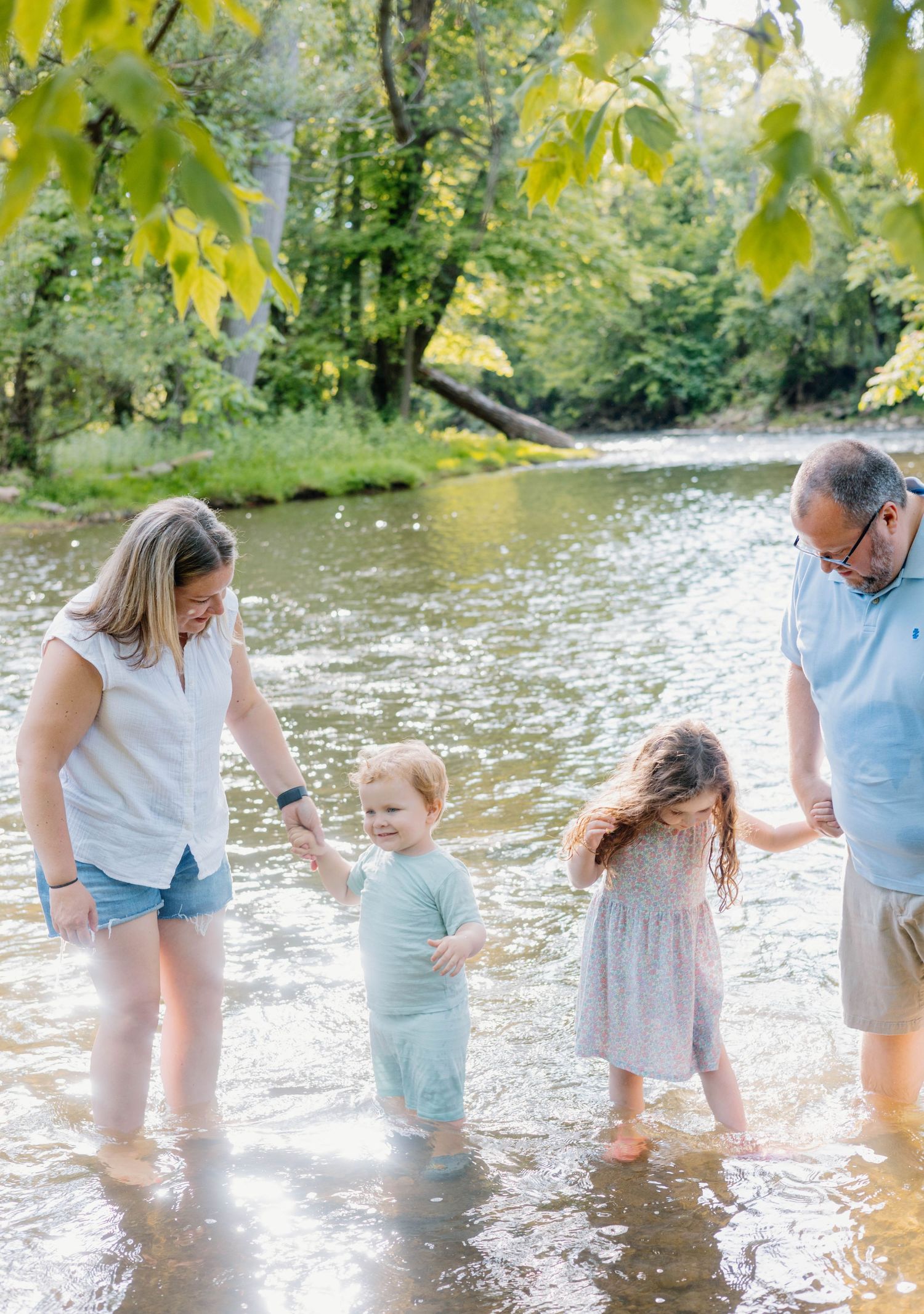 Family of four wading in a shallow creek surrounded by lush green trees on a sunny day.